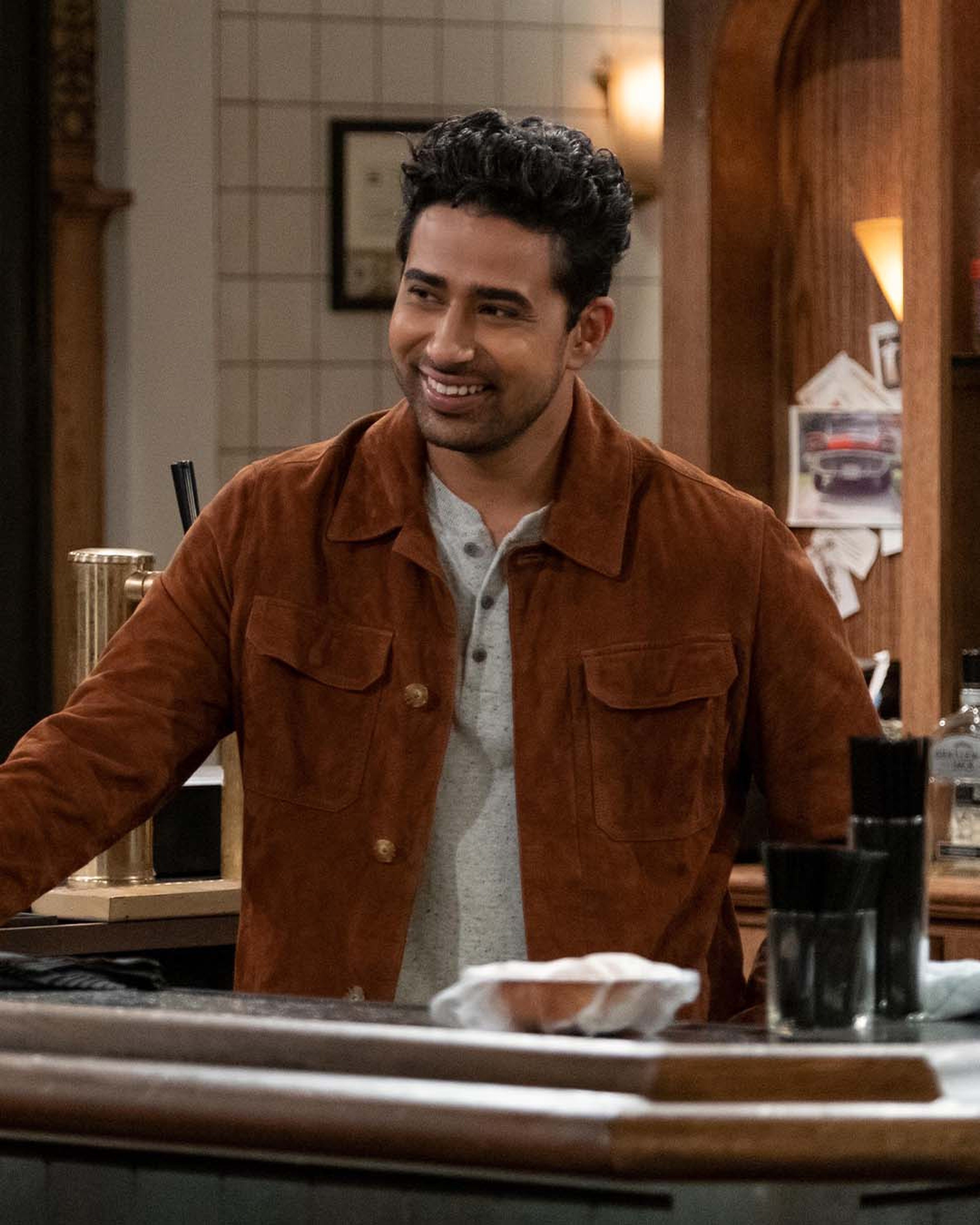 A man with dark curly hair wearing a brown suede jacket, smiling in a bar or restaurant setting.