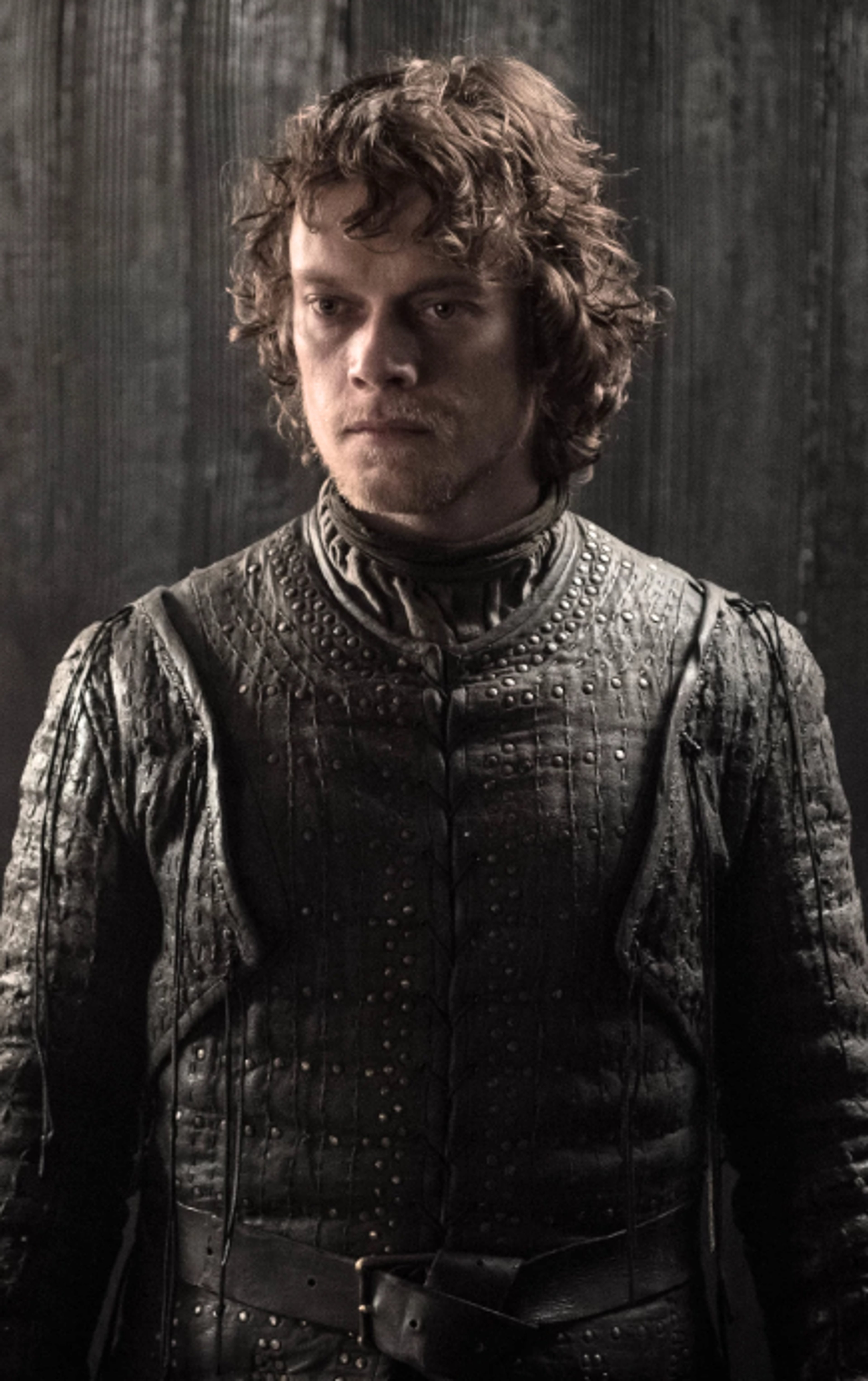 A young man with curly brown hair wearing a dark leather jacket in a dimly lit setting