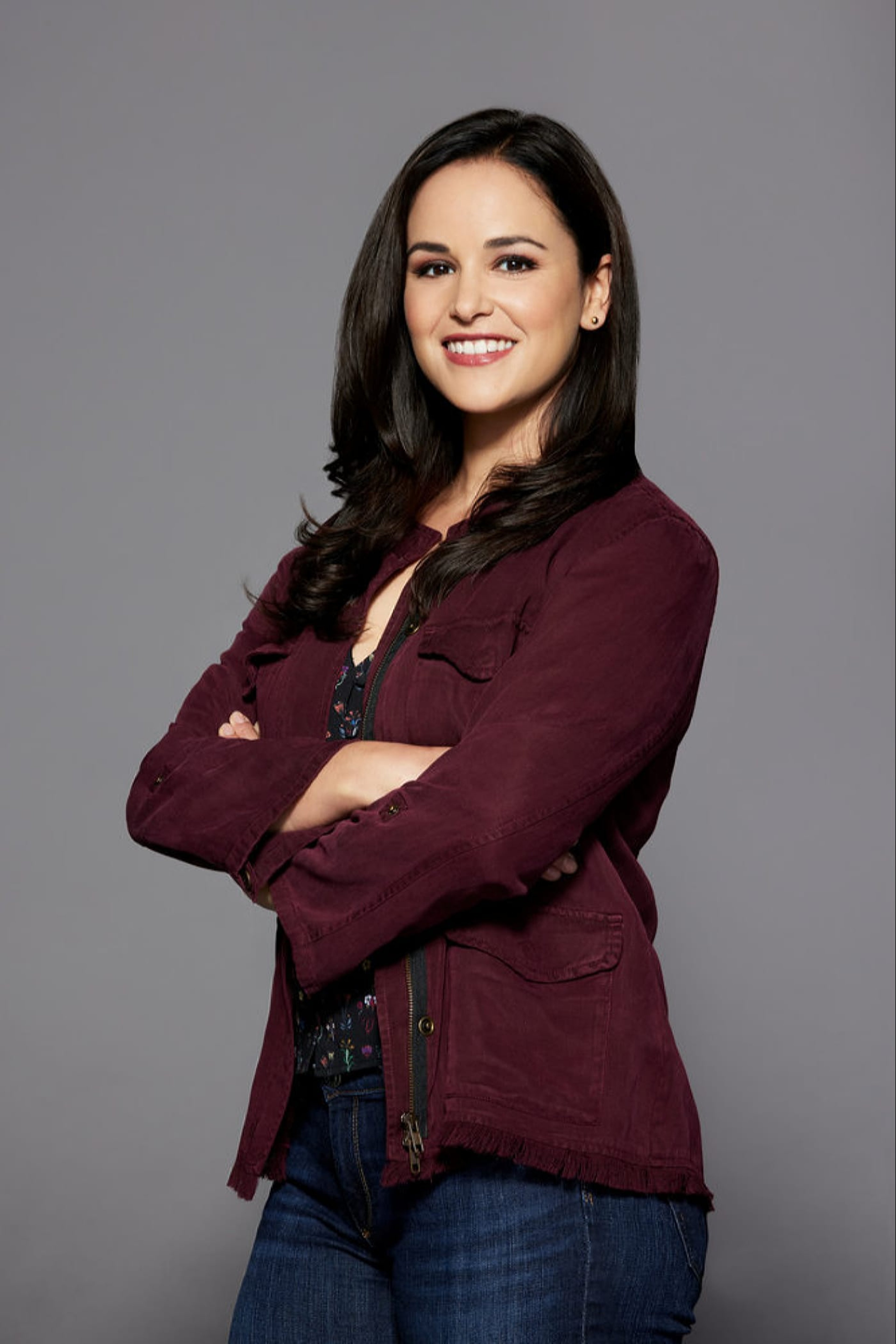 A woman with long dark hair wearing a burgundy jacket, standing with her arms crossed and a friendly expression.