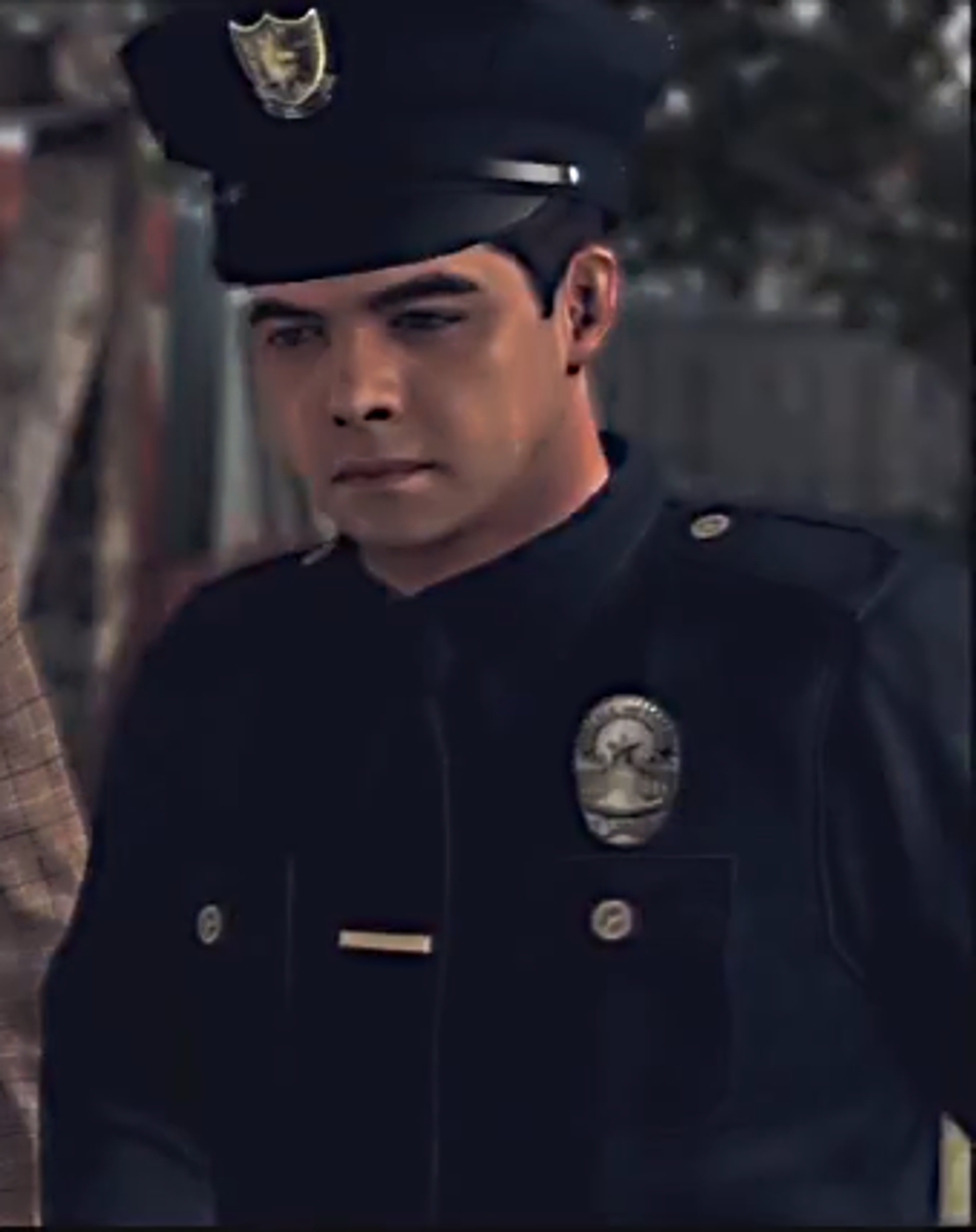 A Hispanic male police officer in a dark uniform standing in a dimly lit urban setting