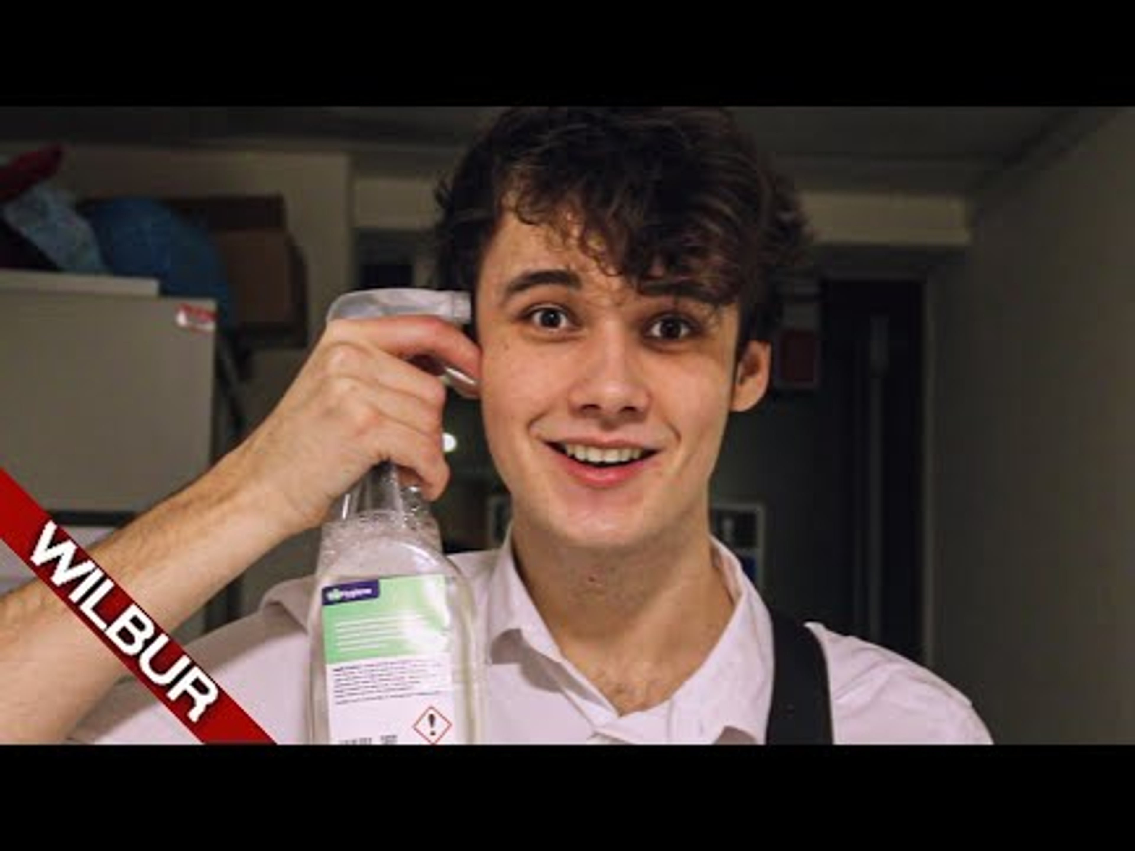 A young man with curly brown hair holding a bottle and smiling