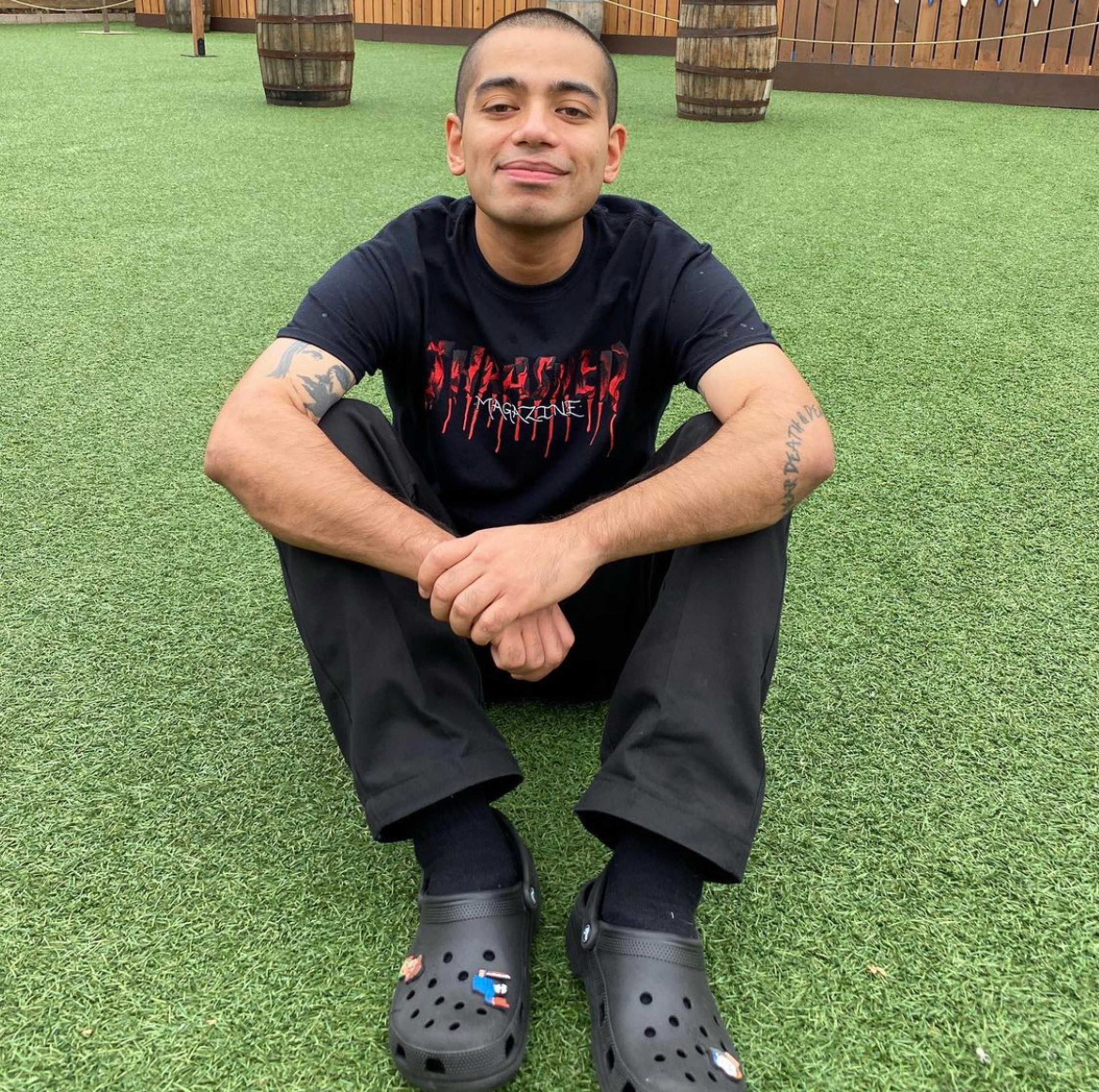 A young man with short dark hair and a beard, wearing a black t-shirt, sitting on a grassy surface with wooden barrels in the background.