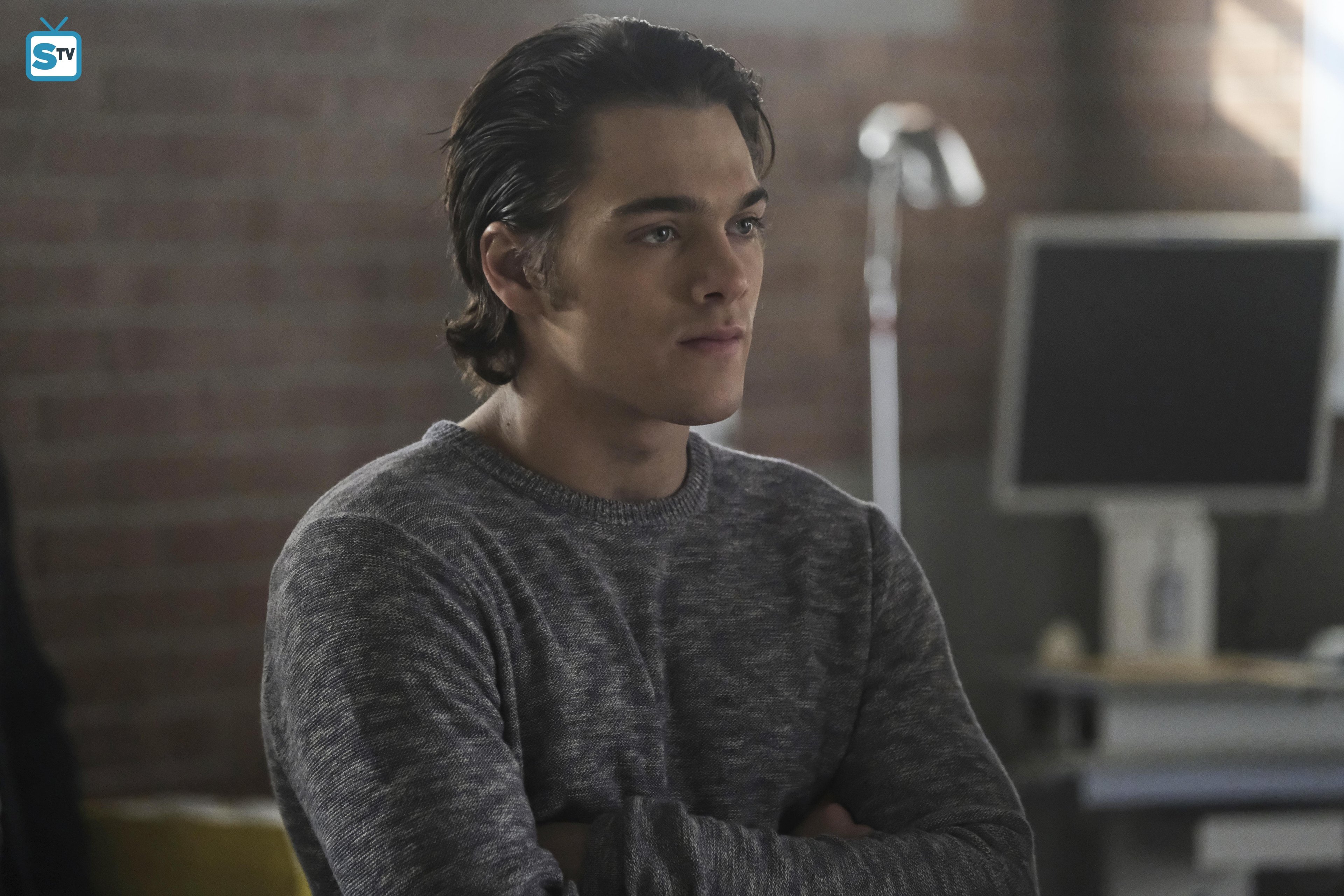 A young man with short dark hair wearing a gray sweater in a dimly lit setting.