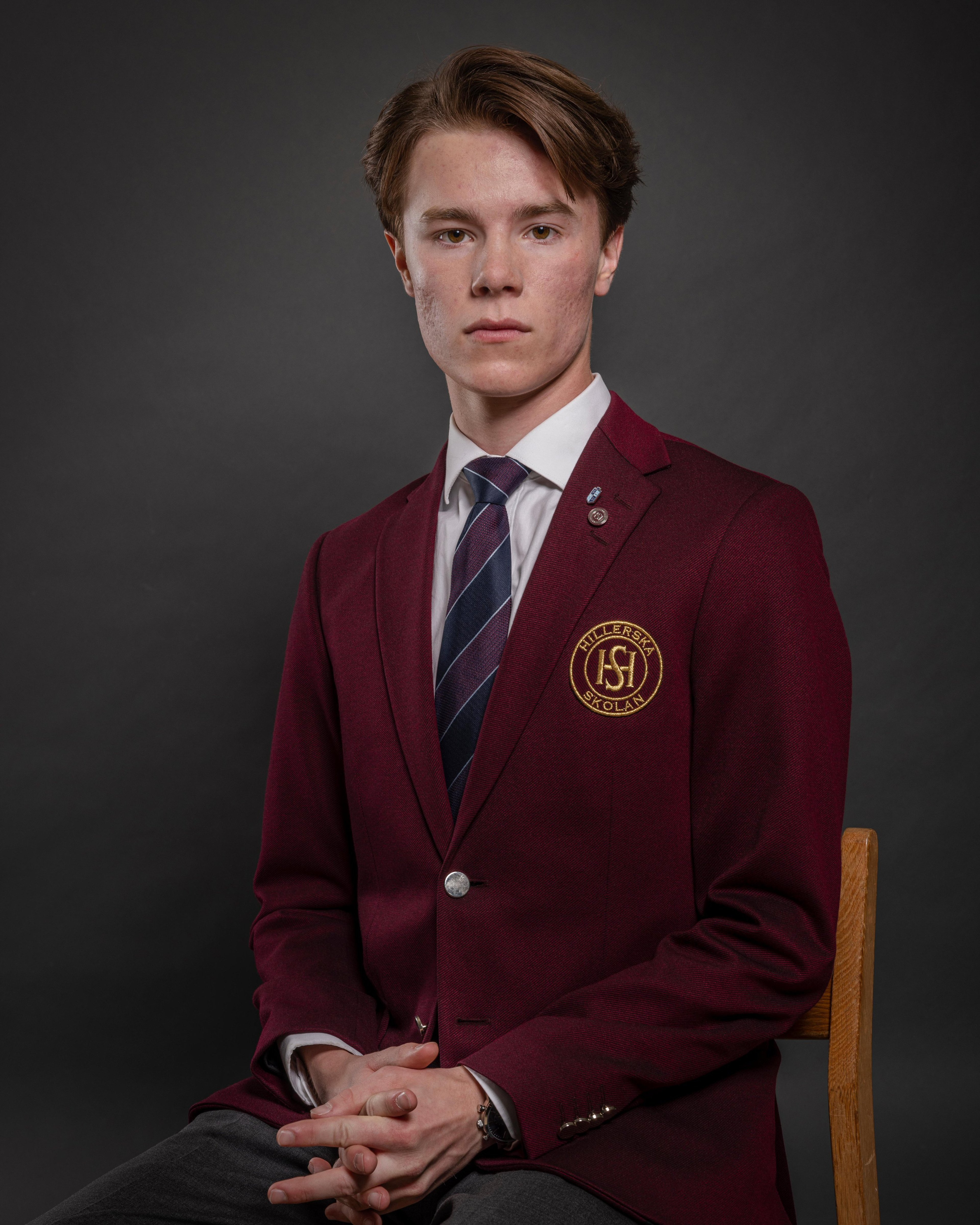 A young man in a burgundy school uniform sitting in a chair against a dark background.