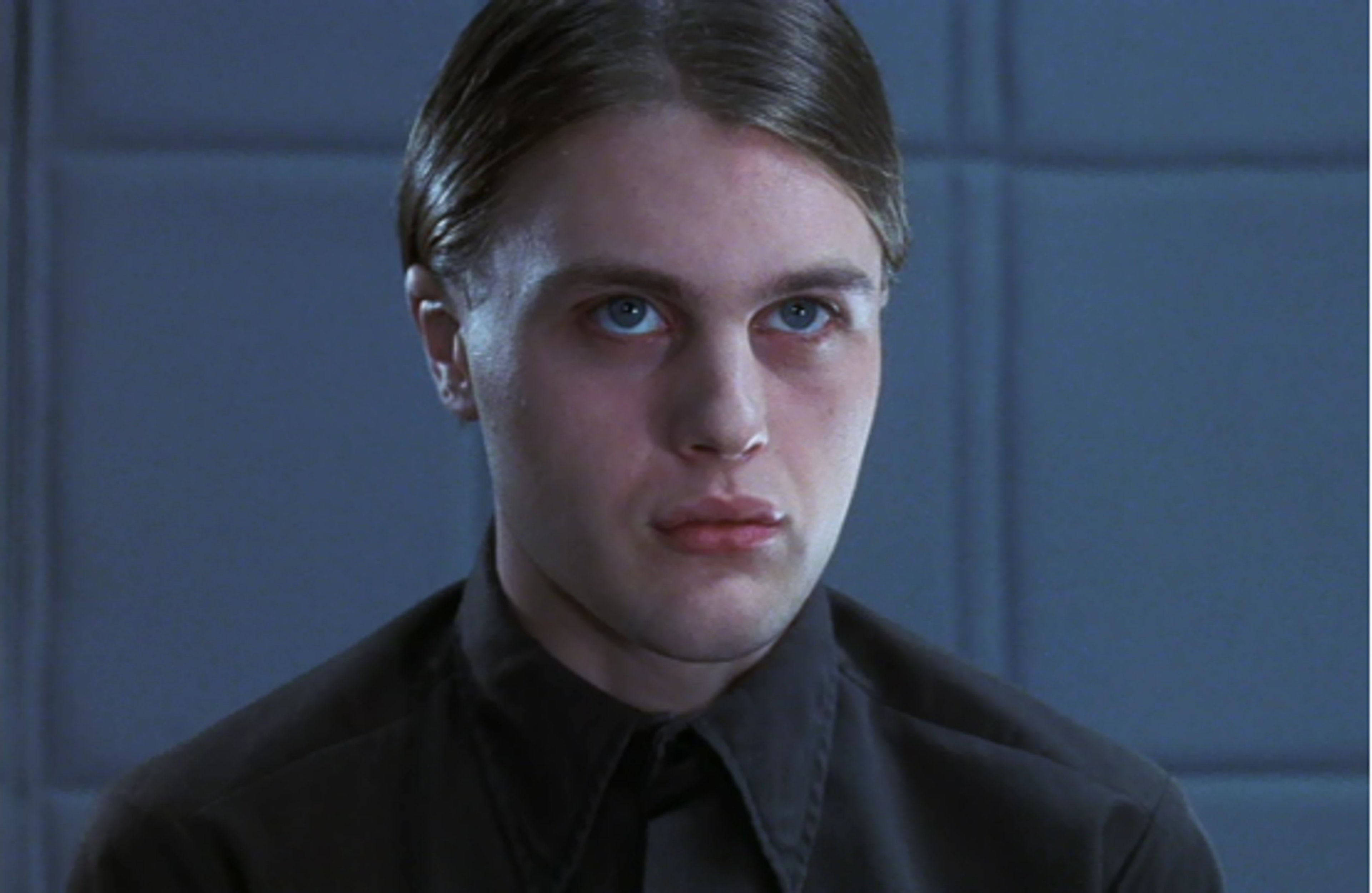 A young man with short dark hair wearing a black shirt in a dimly lit setting