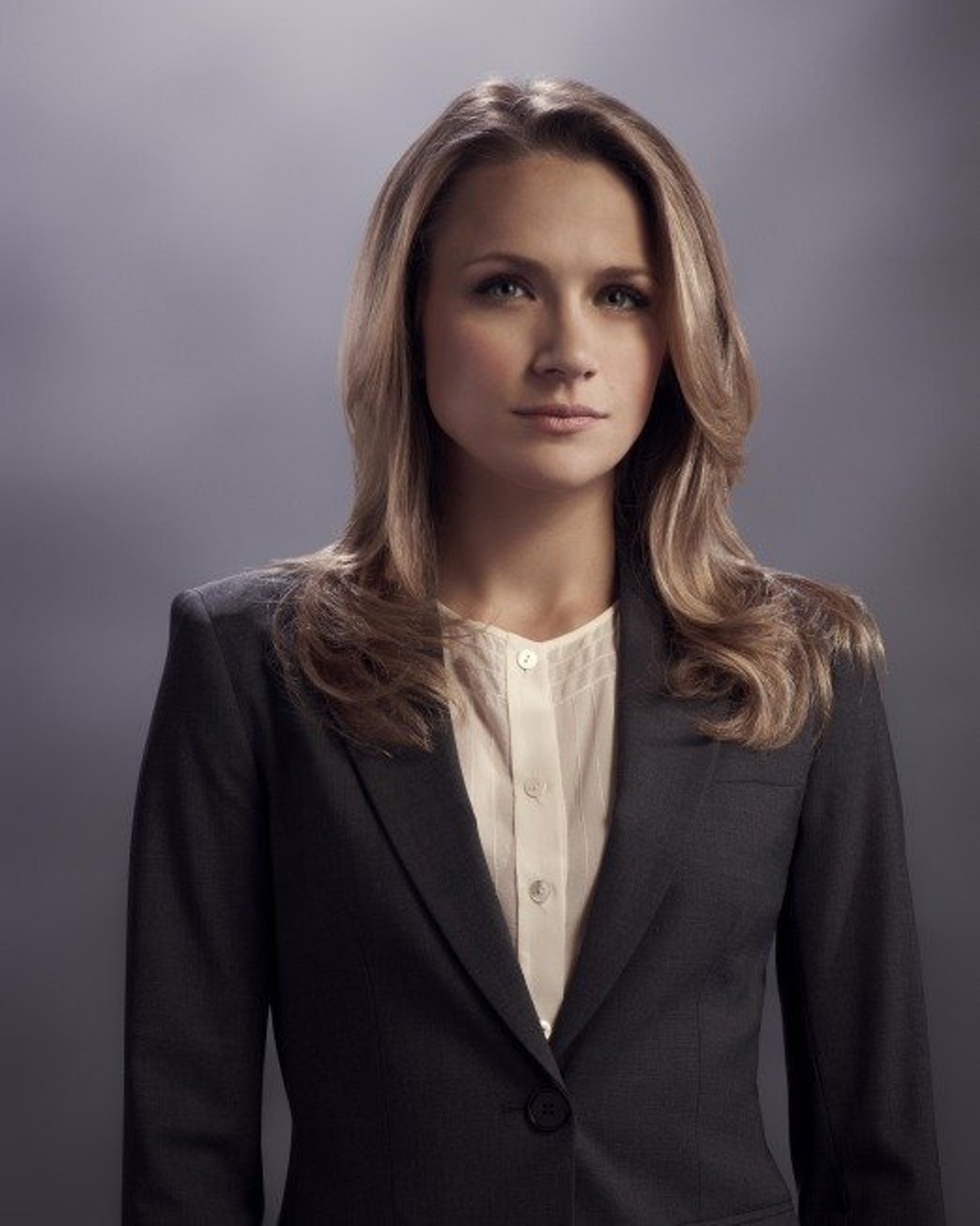 A woman with long brown hair wearing a black blazer and white blouse, looking serious and determined.