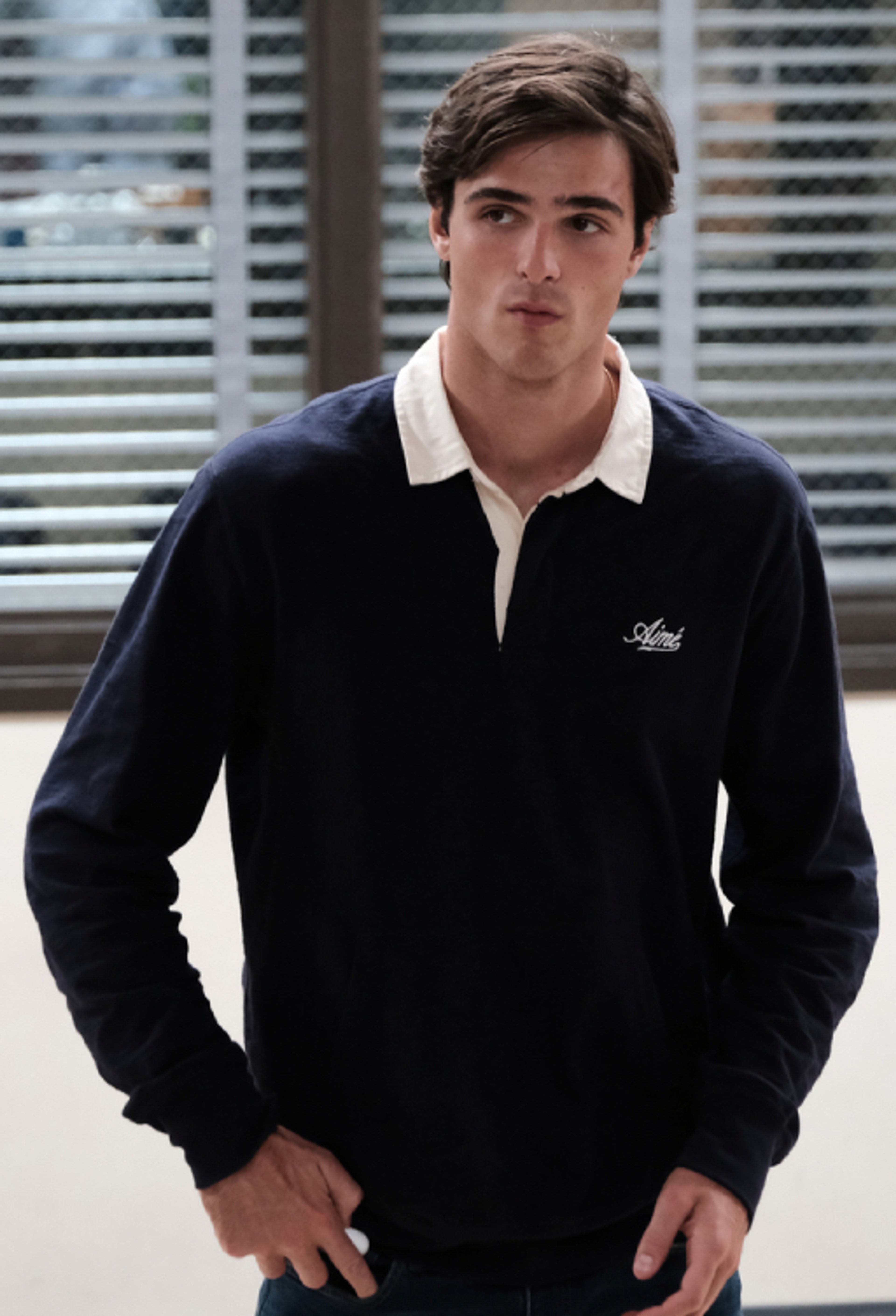 A young man with dark hair and eyes, wearing a black sweater with a white collar, standing in front of a blurred background.