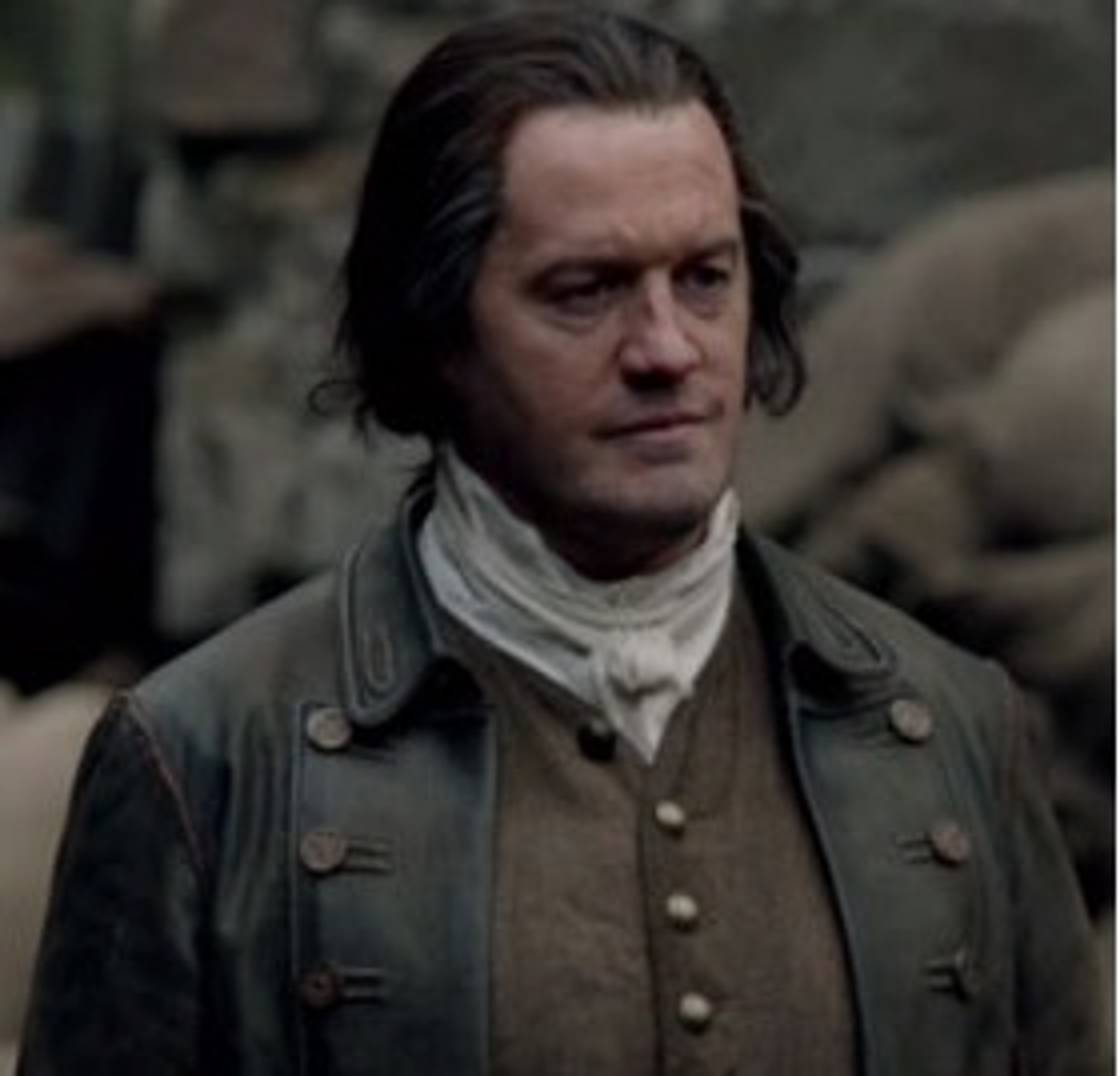 A man in a military-style coat standing in a rural outdoor setting