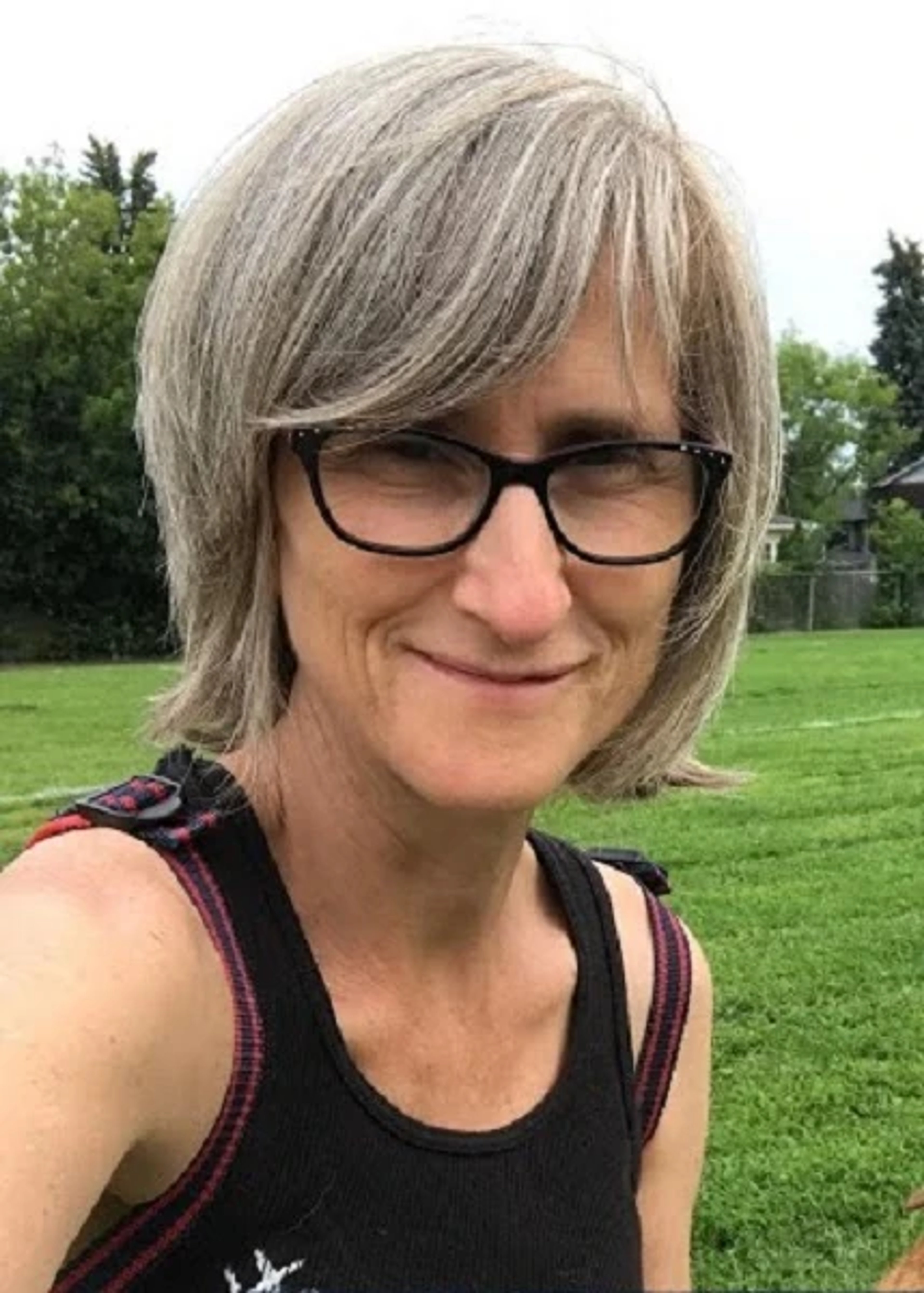 A middle-aged woman with short grey-white hair wearing a black tank top, standing in a grassy outdoor area.