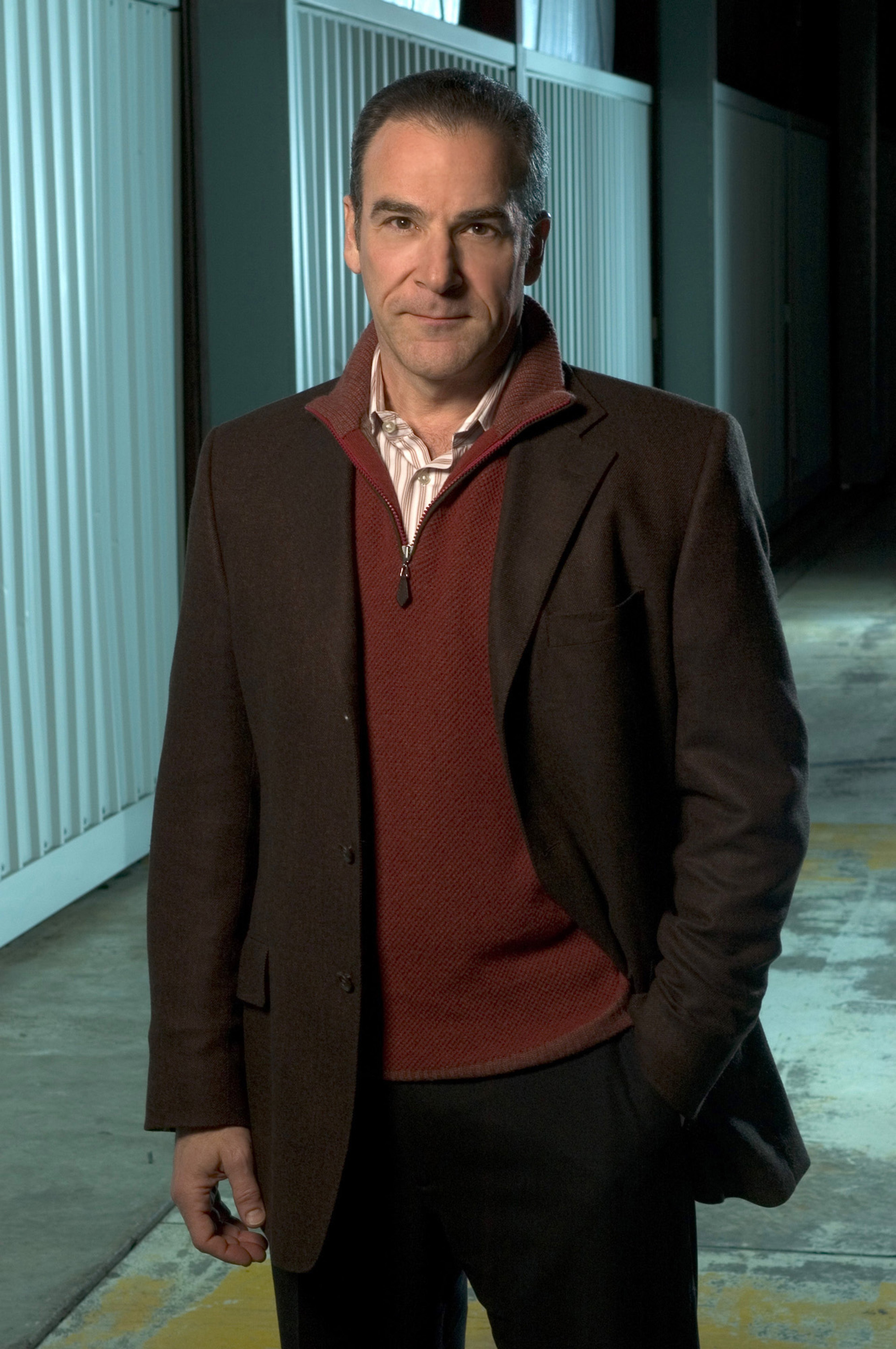 A middle-aged man with gray hair and a beard, wearing a red sweater and a brown coat, standing in a hallway or corridor.