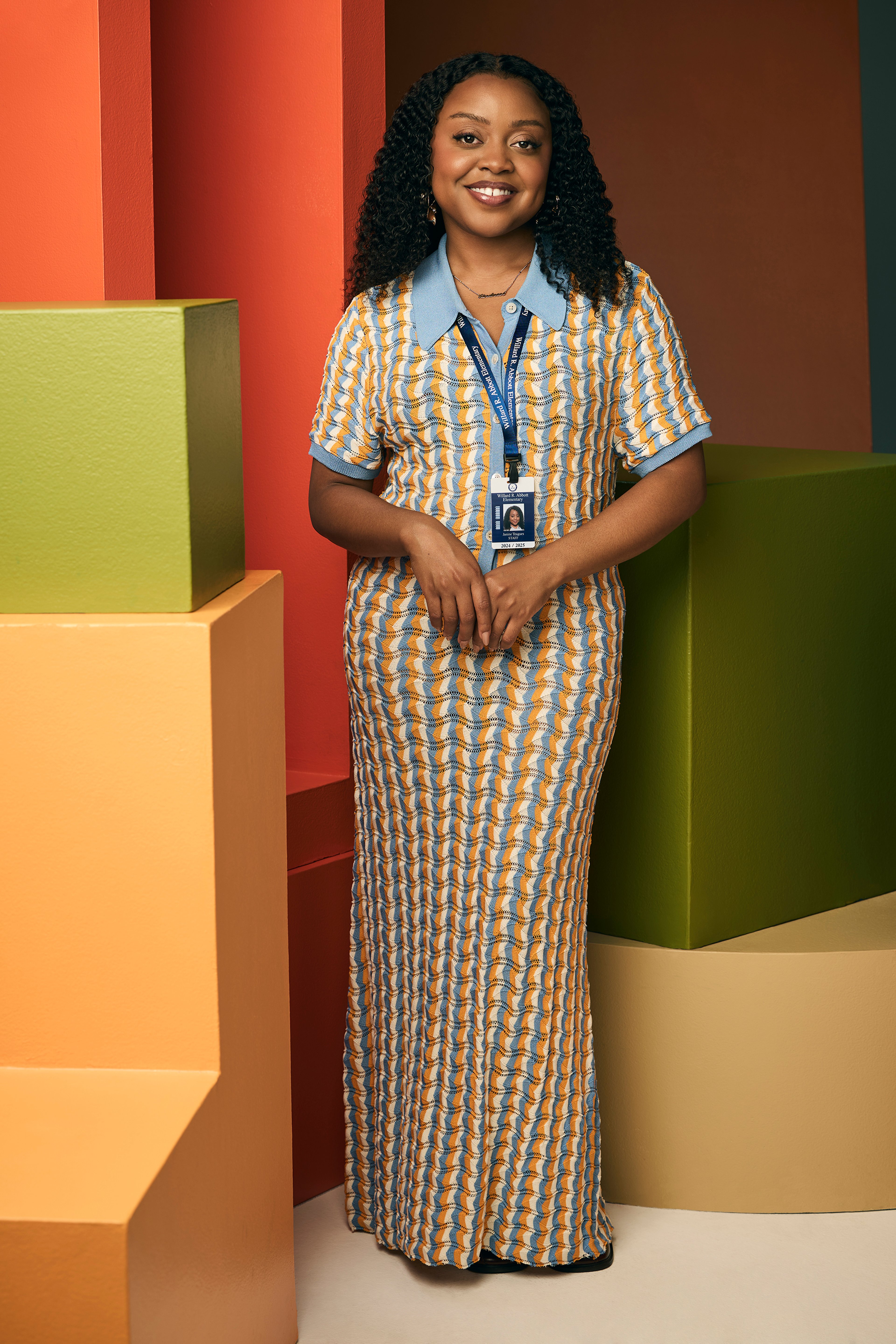 A young Black woman with curly hair wearing a yellow and white checkered dress, standing in front of a colorful background.