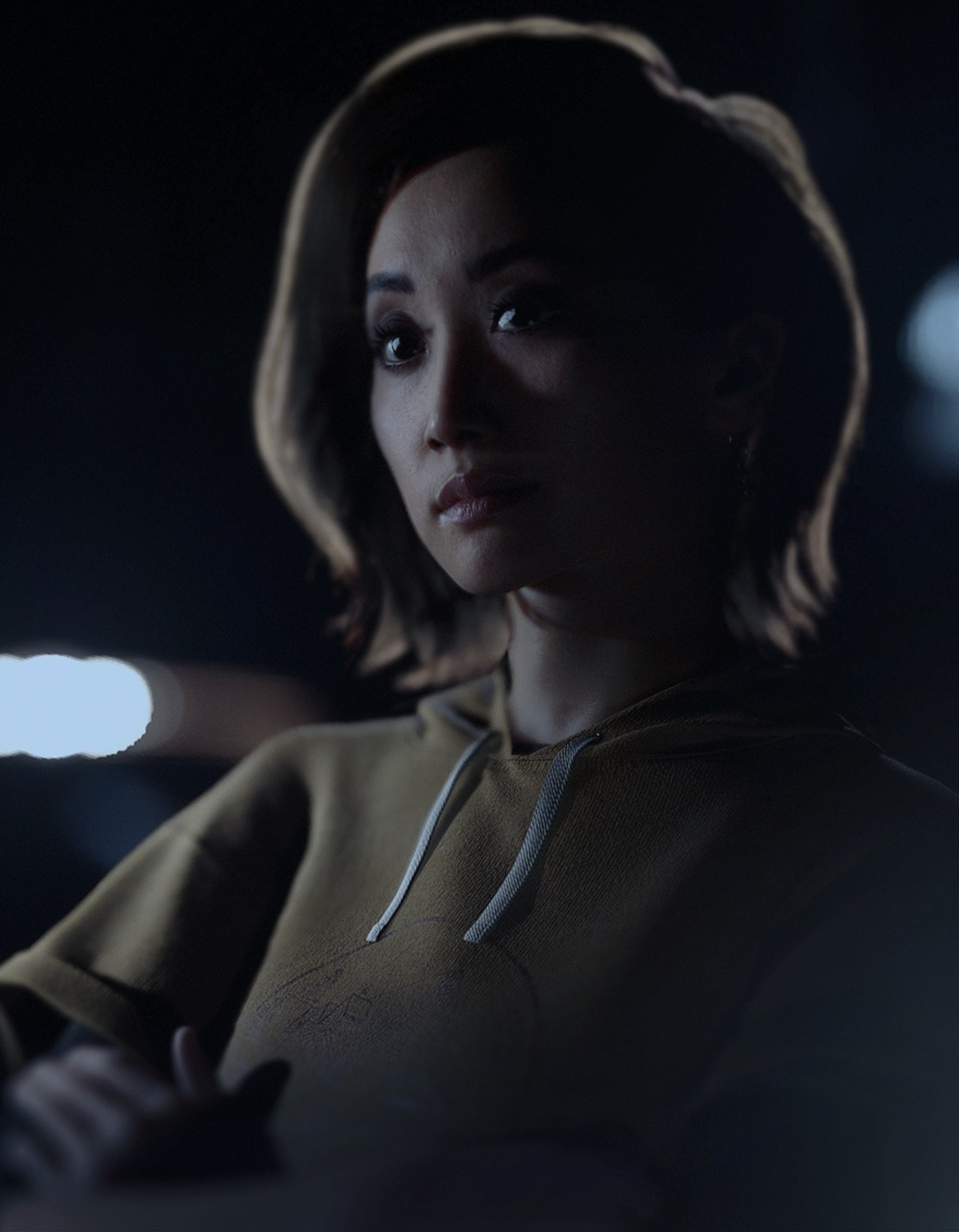 A young woman with dark hair in a serious expression, wearing a light-colored jacket or sweater in a dimly lit environment.