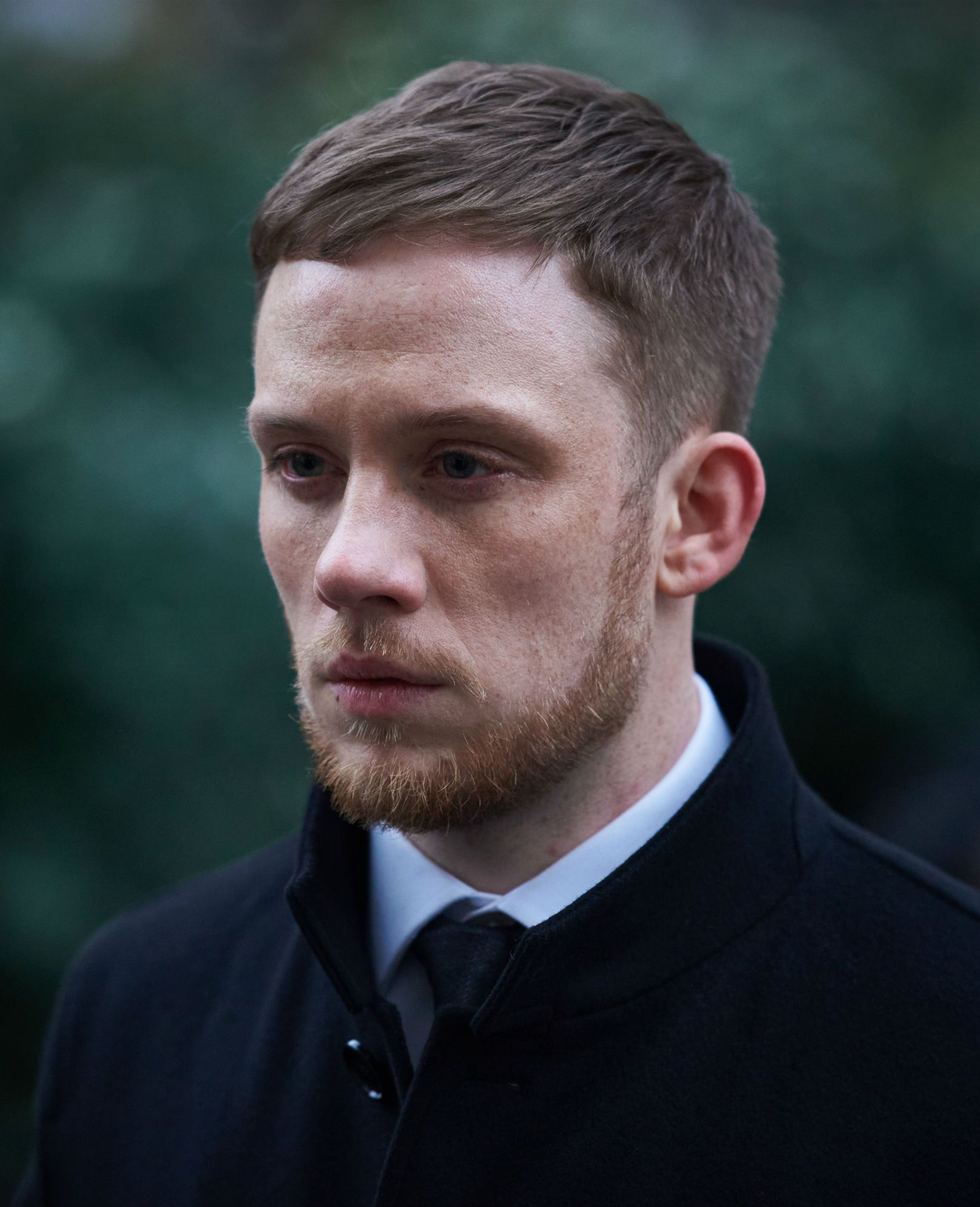 A man with short brown hair and a beard, wearing a dark suit, with a serious expression on his face.