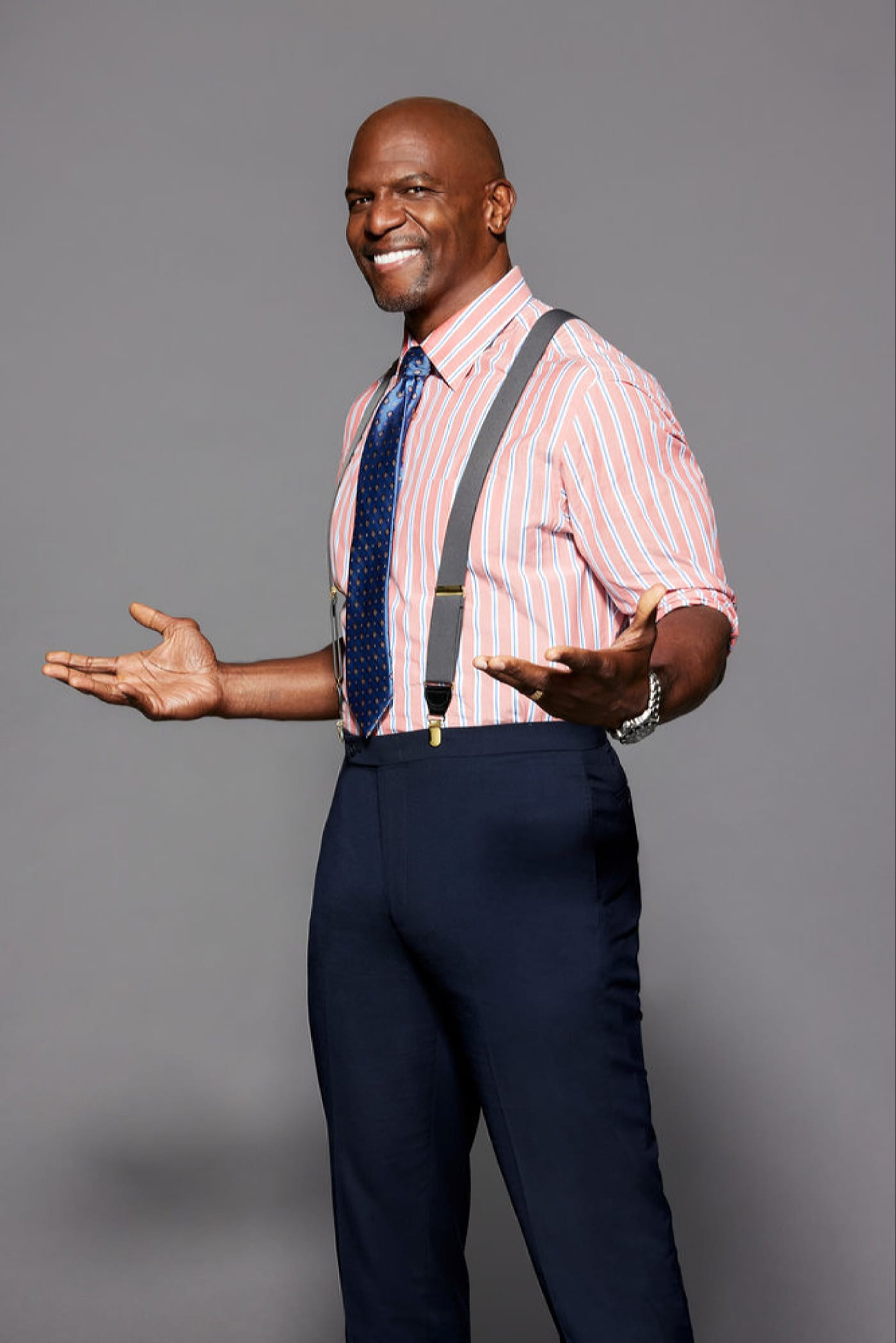 A smiling, muscular Black man in his 50s wearing a pink and white striped shirt, navy blue suspenders, and pants.