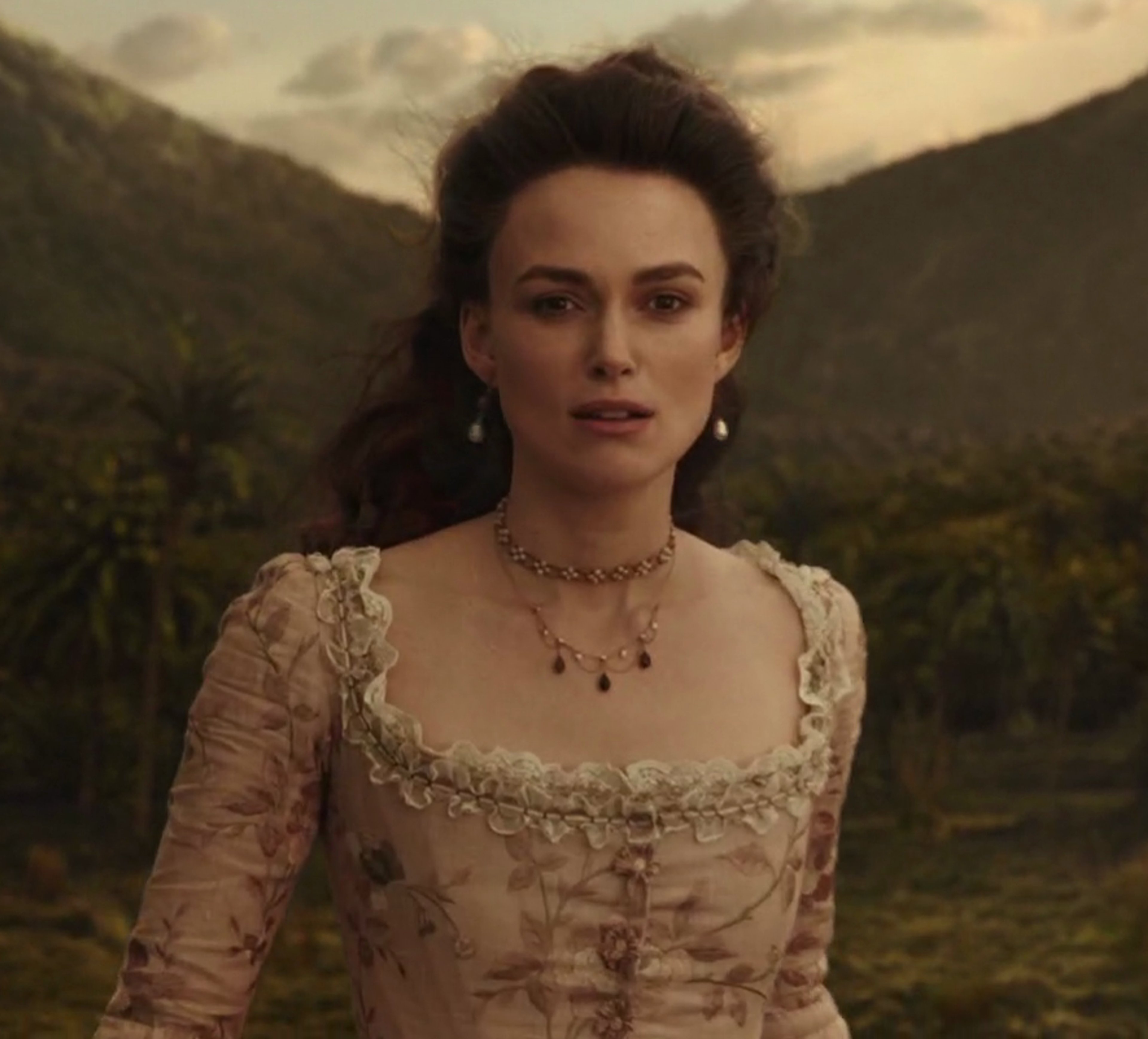 A young woman with dark curly hair wearing a lace dress, standing in a scenic outdoor setting.