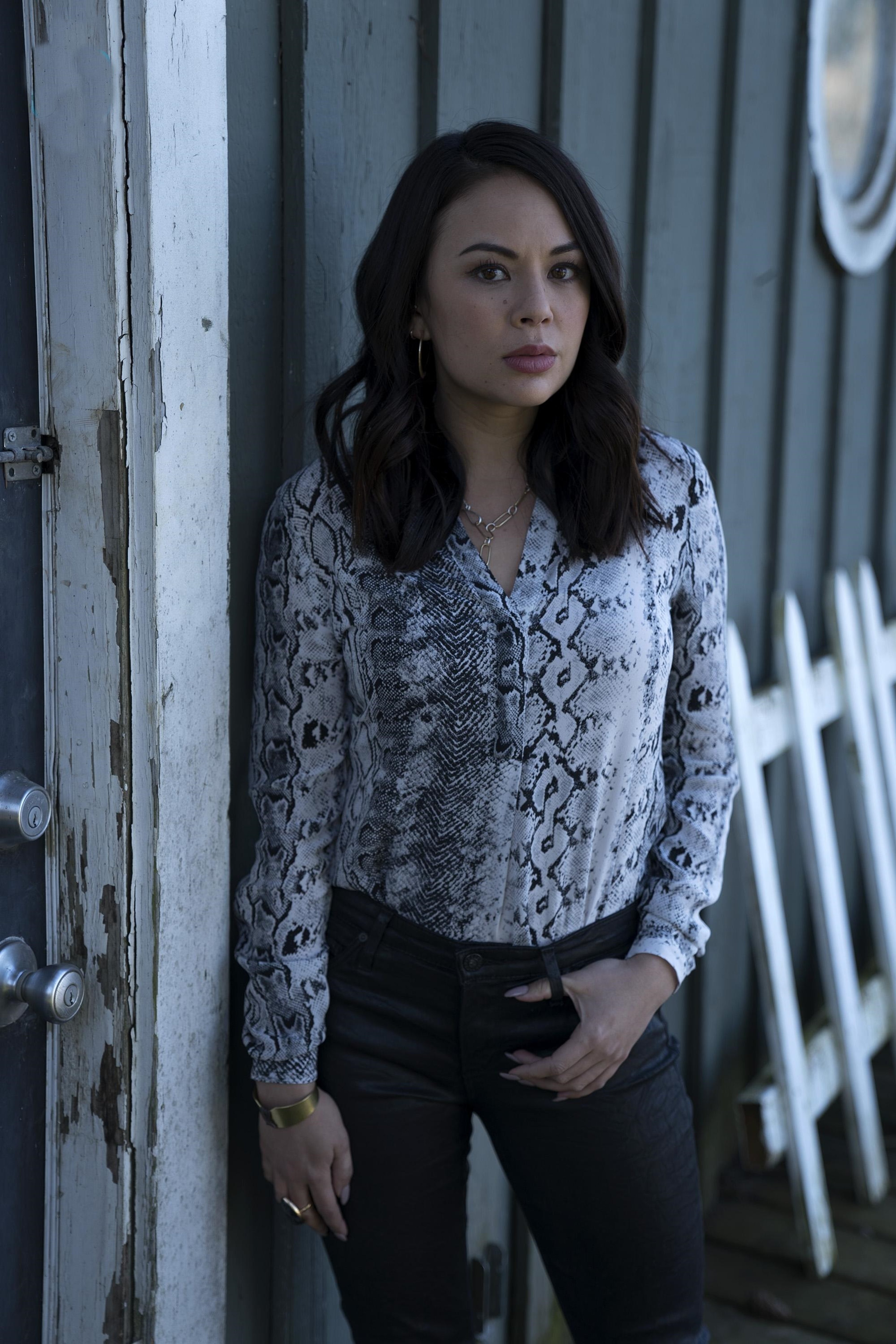 A young woman with dark hair wearing a patterned blouse, standing in front of a wooden door or wall.