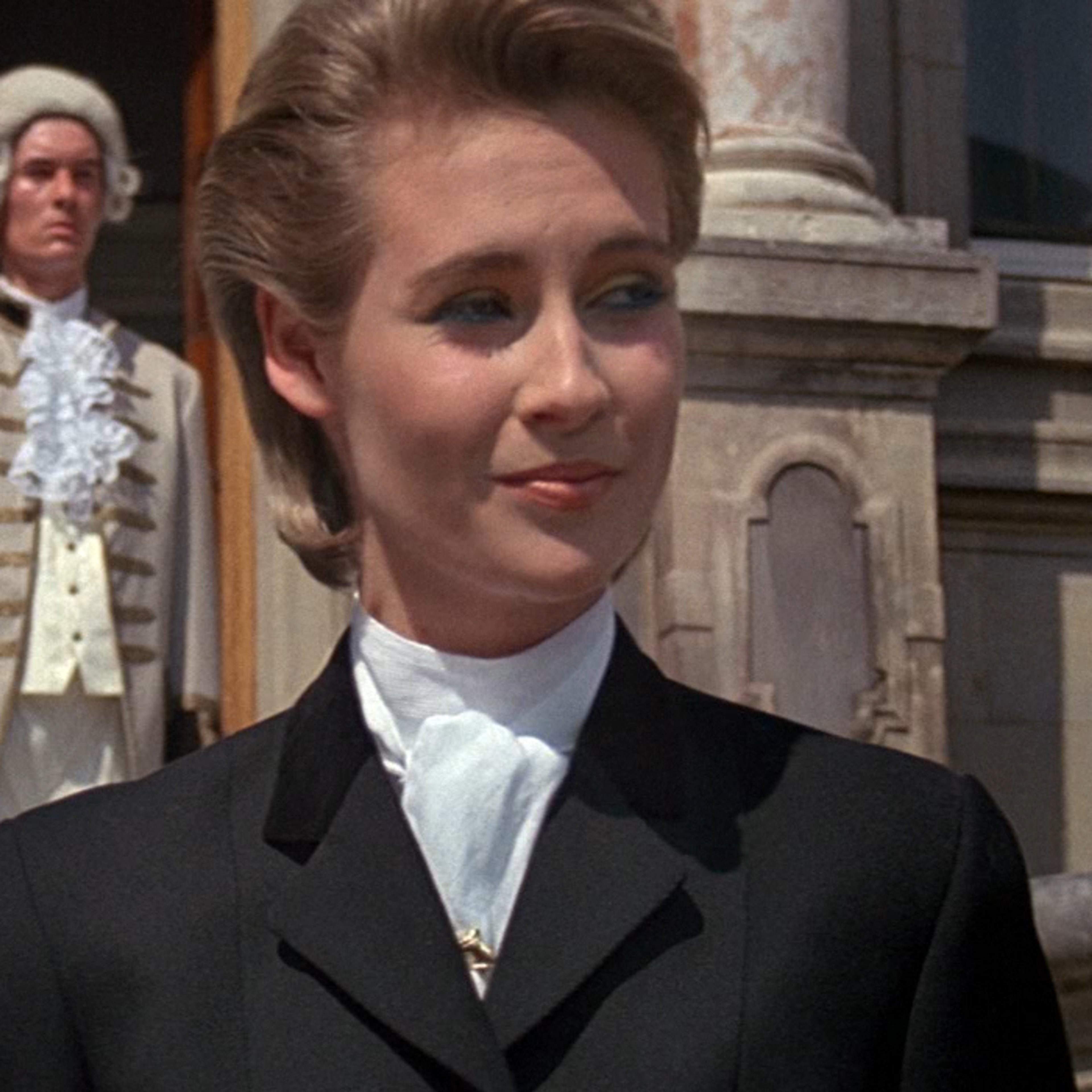 A woman in a black suit jacket and white shirt with an updo hairstyle, standing in a formal setting.