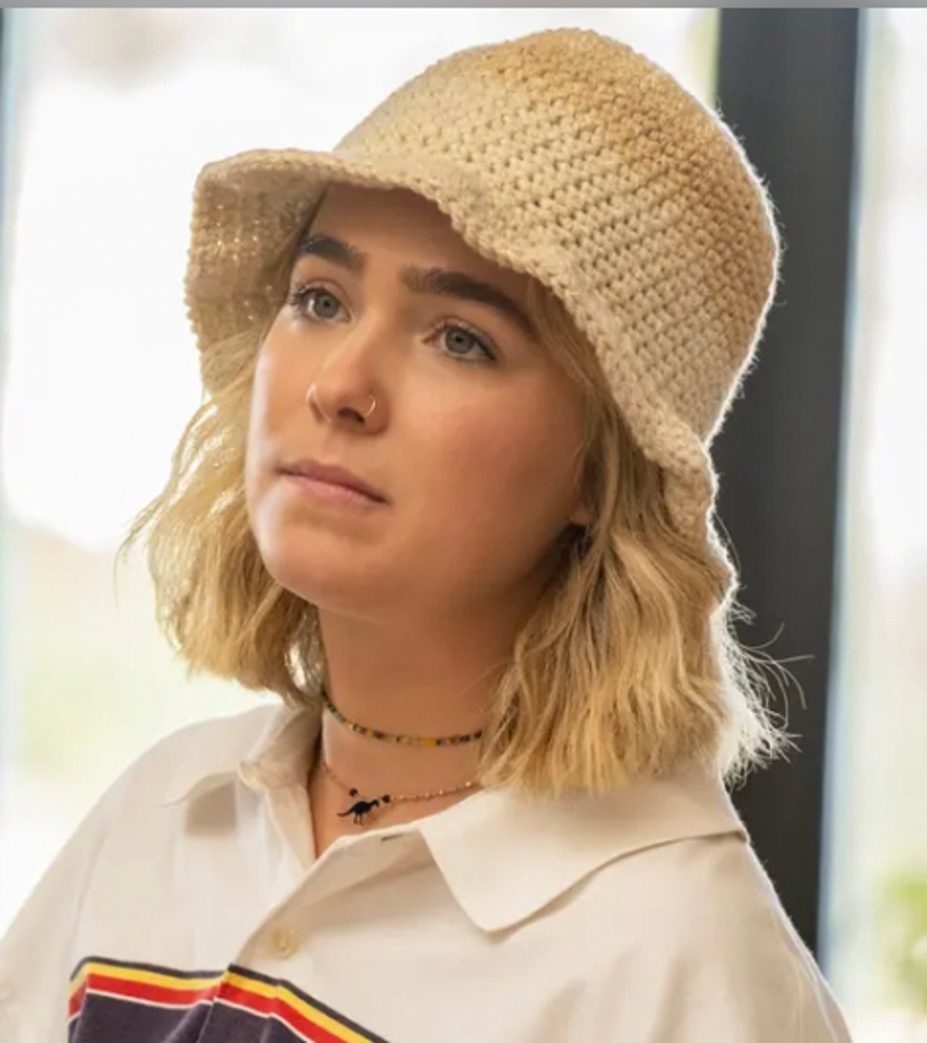 A young woman with blonde hair wearing a beige crocheted hat and a white collared shirt with a striped pattern.