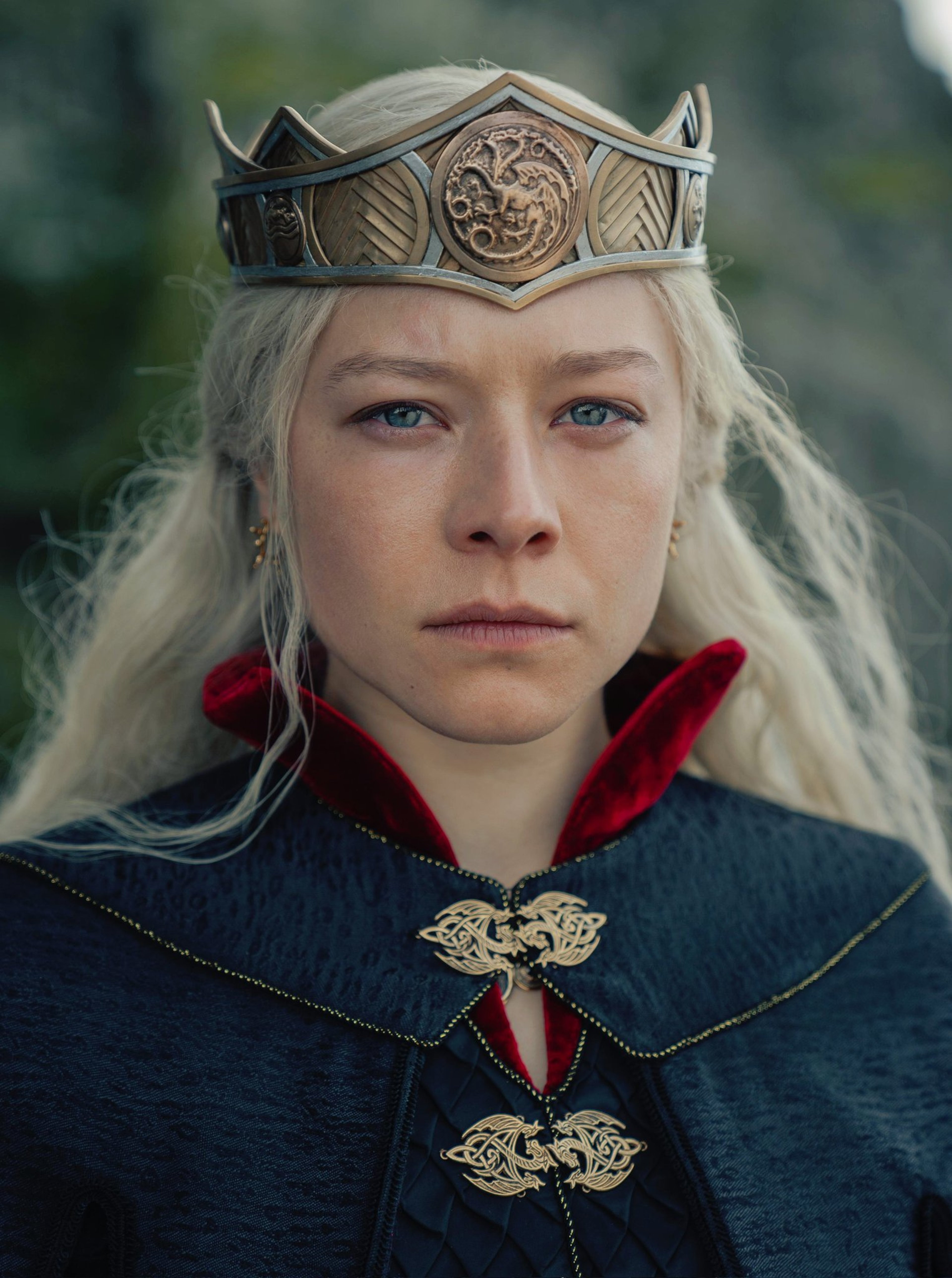 A young woman with blonde hair wearing a crown-like headpiece and a dark cloak, standing in an outdoor setting.