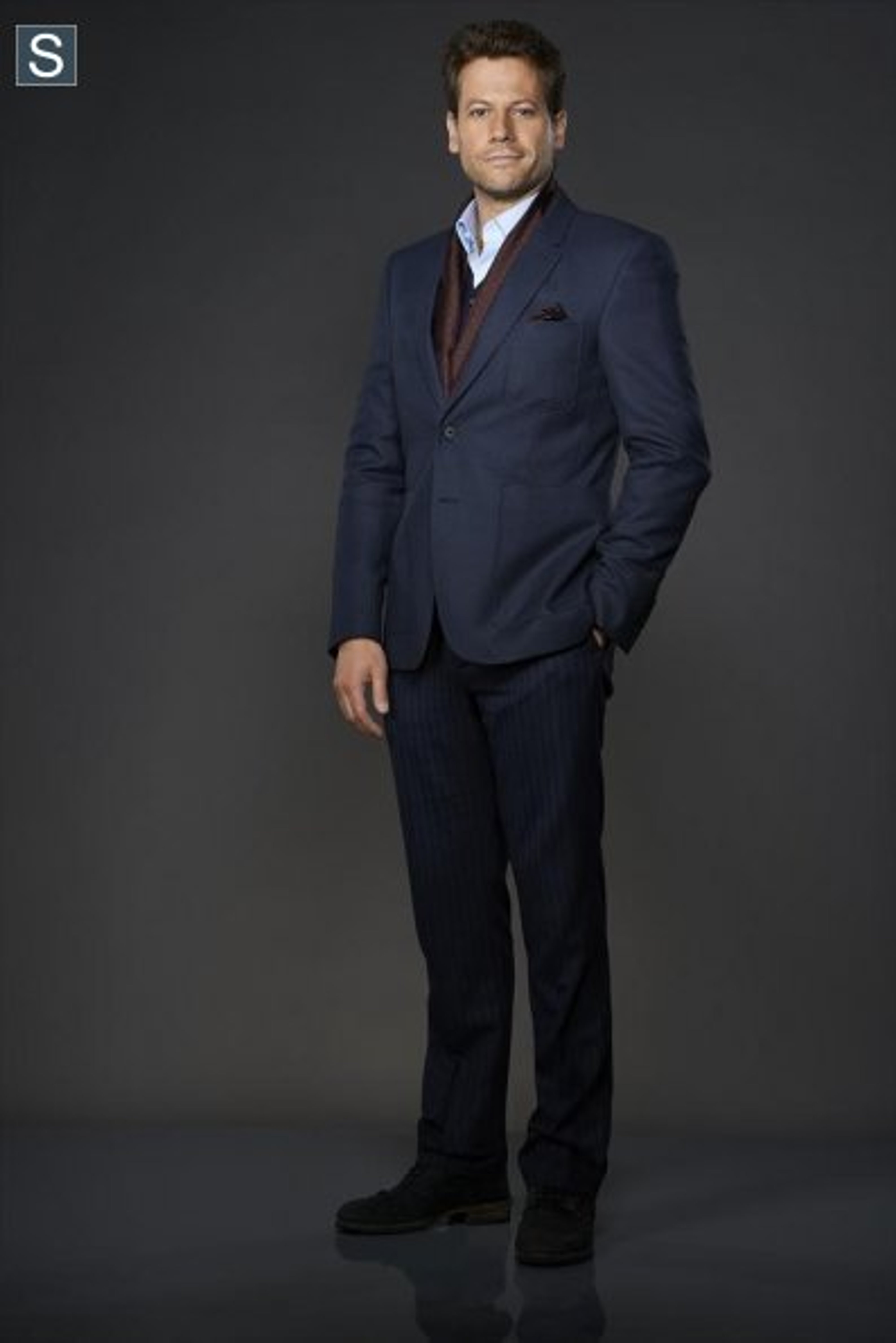 A man in a navy blue suit and burgundy tie standing in a dark studio setting.