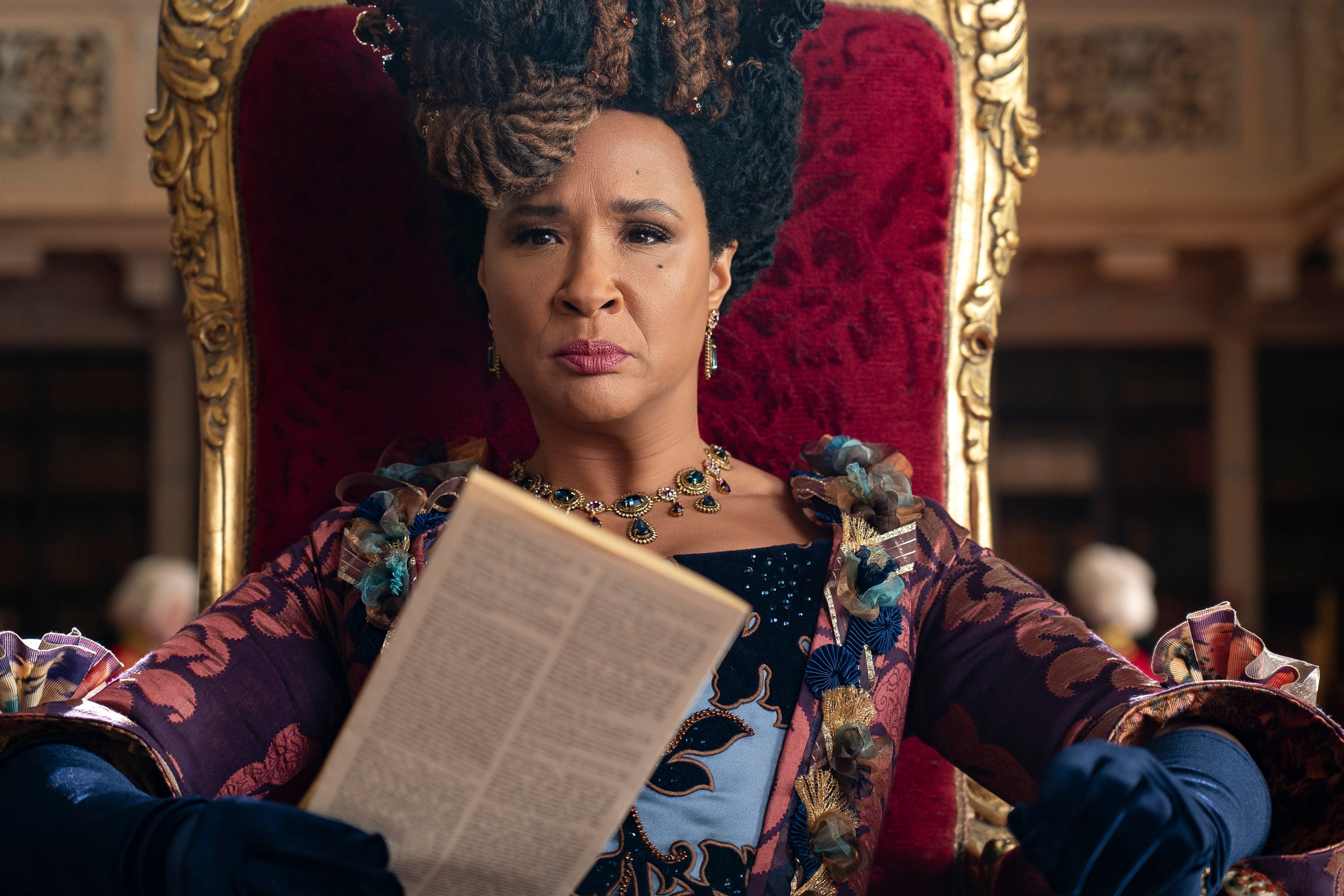 A woman in an elaborate dress and crown, seated in a throne-like chair in a lavish interior setting.