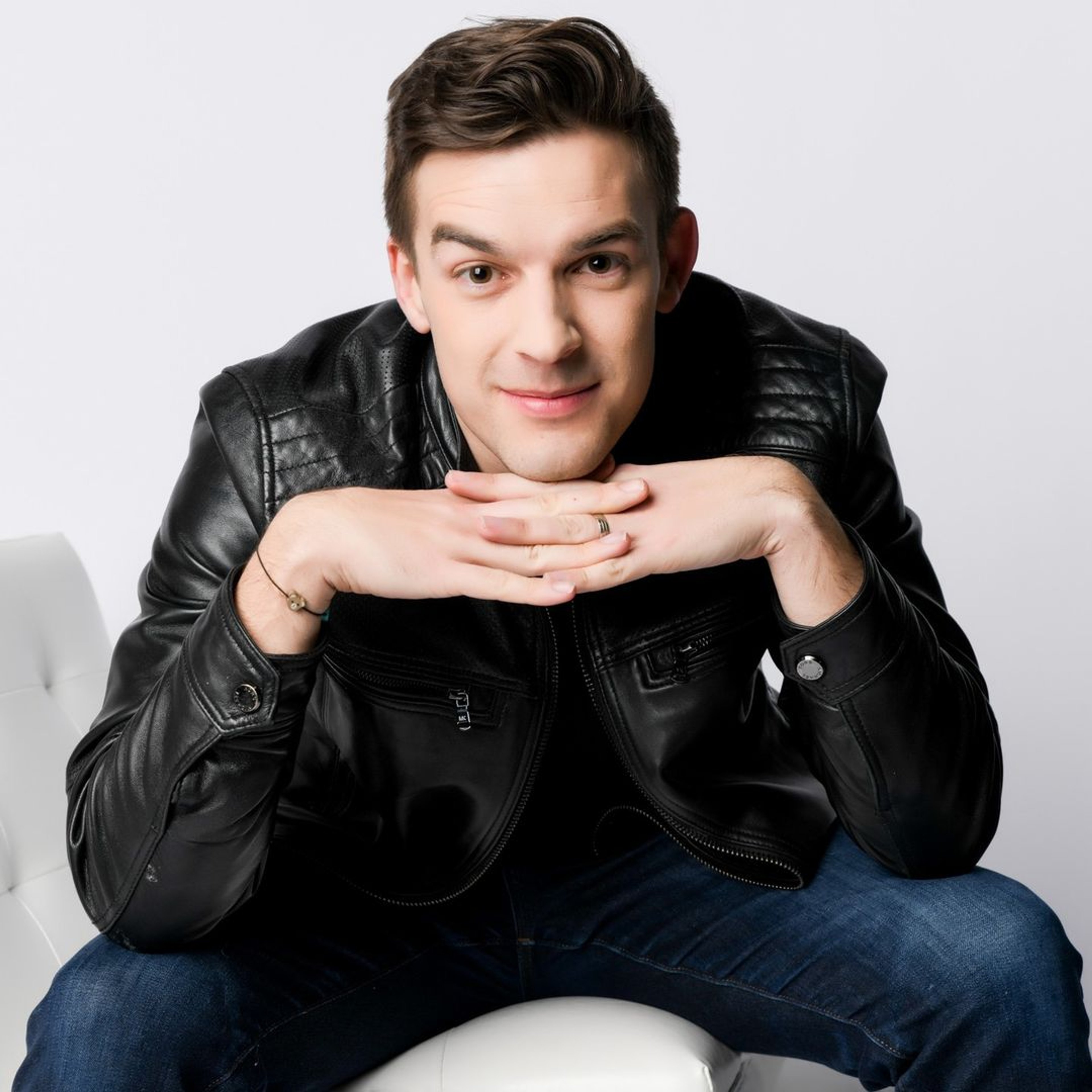 A man with short dark hair wearing a black leather jacket, sitting on a chair and looking at the camera with a friendly expression.