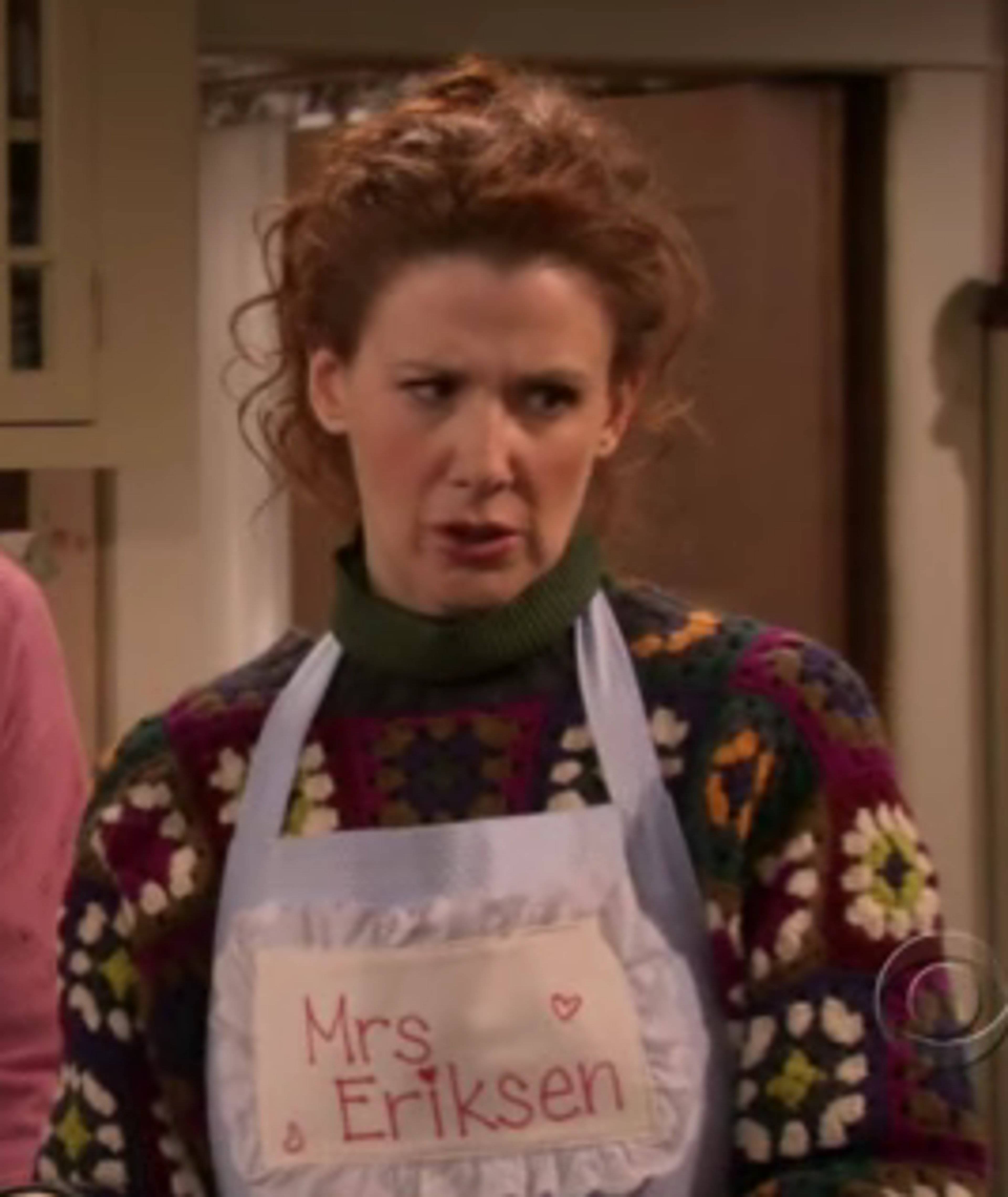 A middle-aged woman with curly red hair wearing a floral apron in a kitchen or home setting.