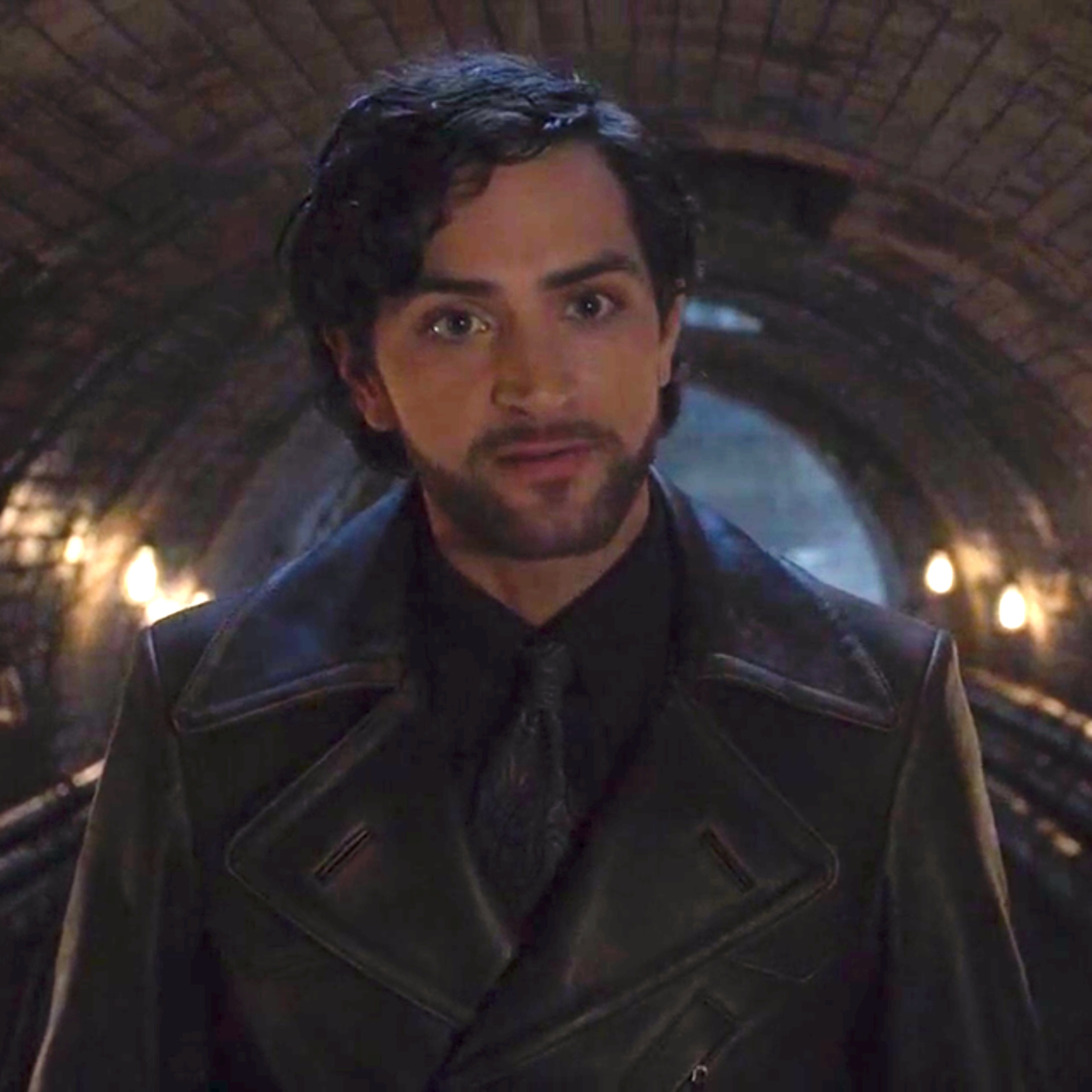 A man with dark hair and a beard wearing a black coat, standing in a dimly lit industrial setting.