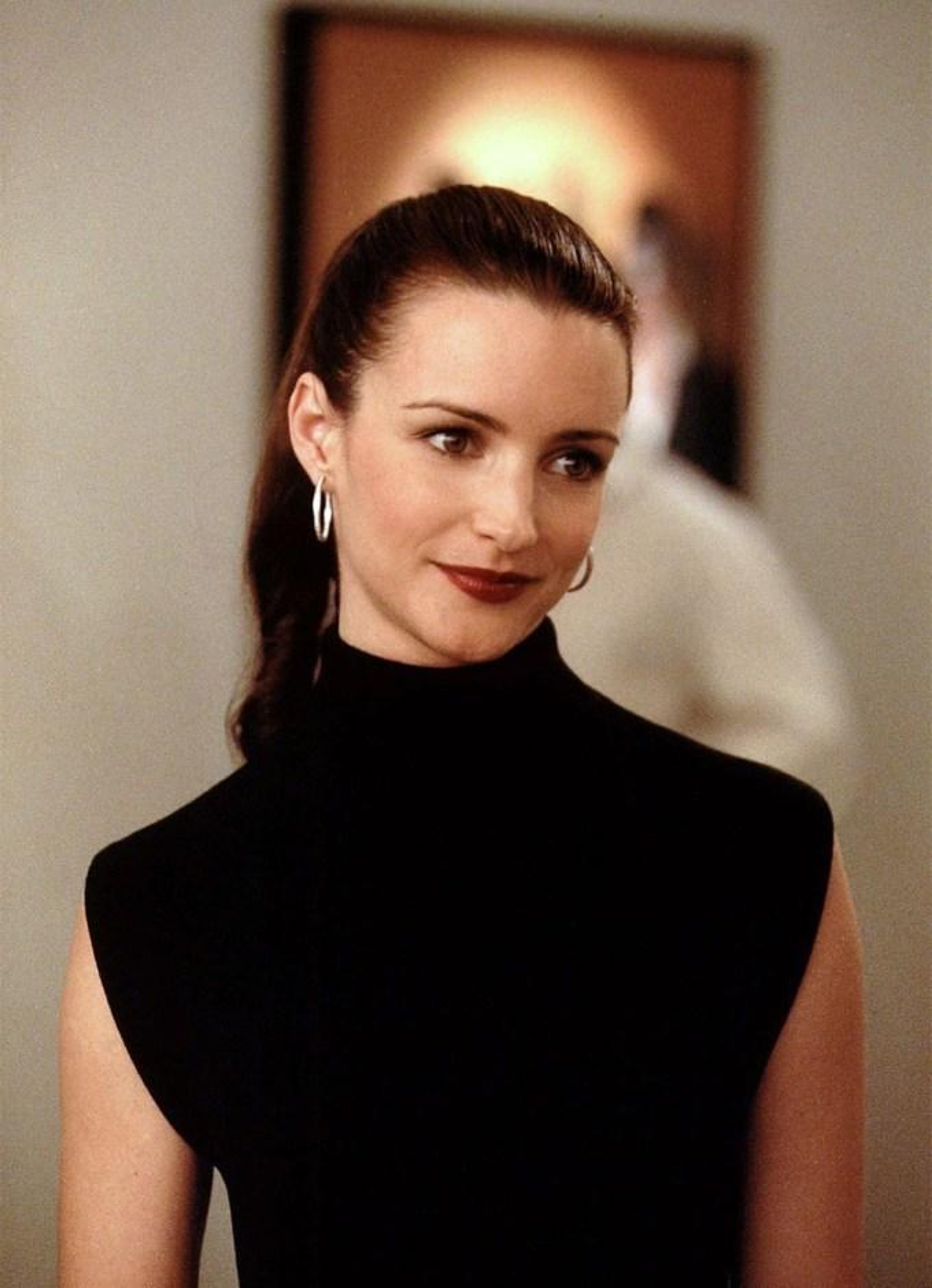 A woman with dark hair in an updo, wearing a black turtleneck dress, looking directly at the camera with a slight smile.