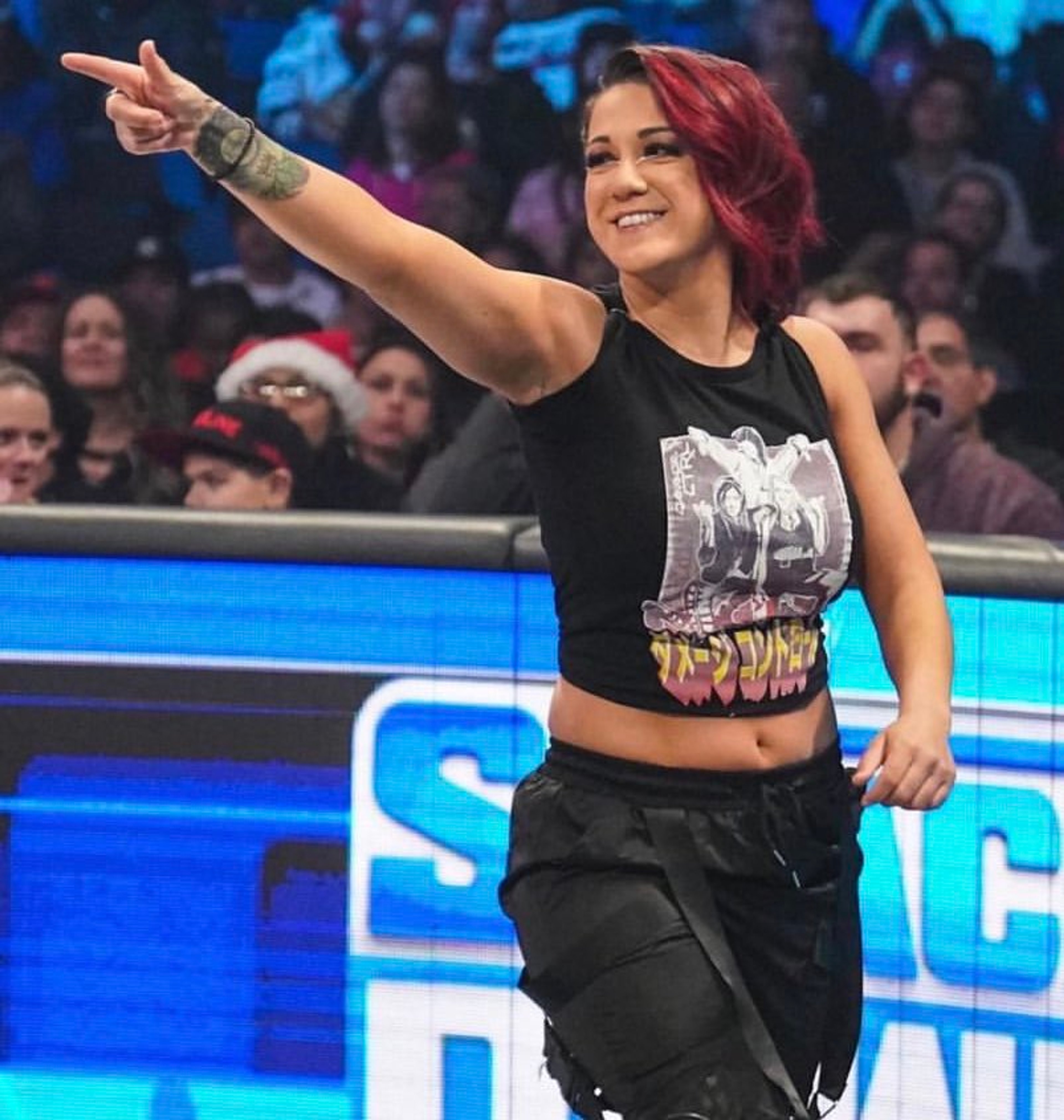 A woman with red hair wearing a black crop top and making a pointing gesture in a wrestling ring or stage setting.