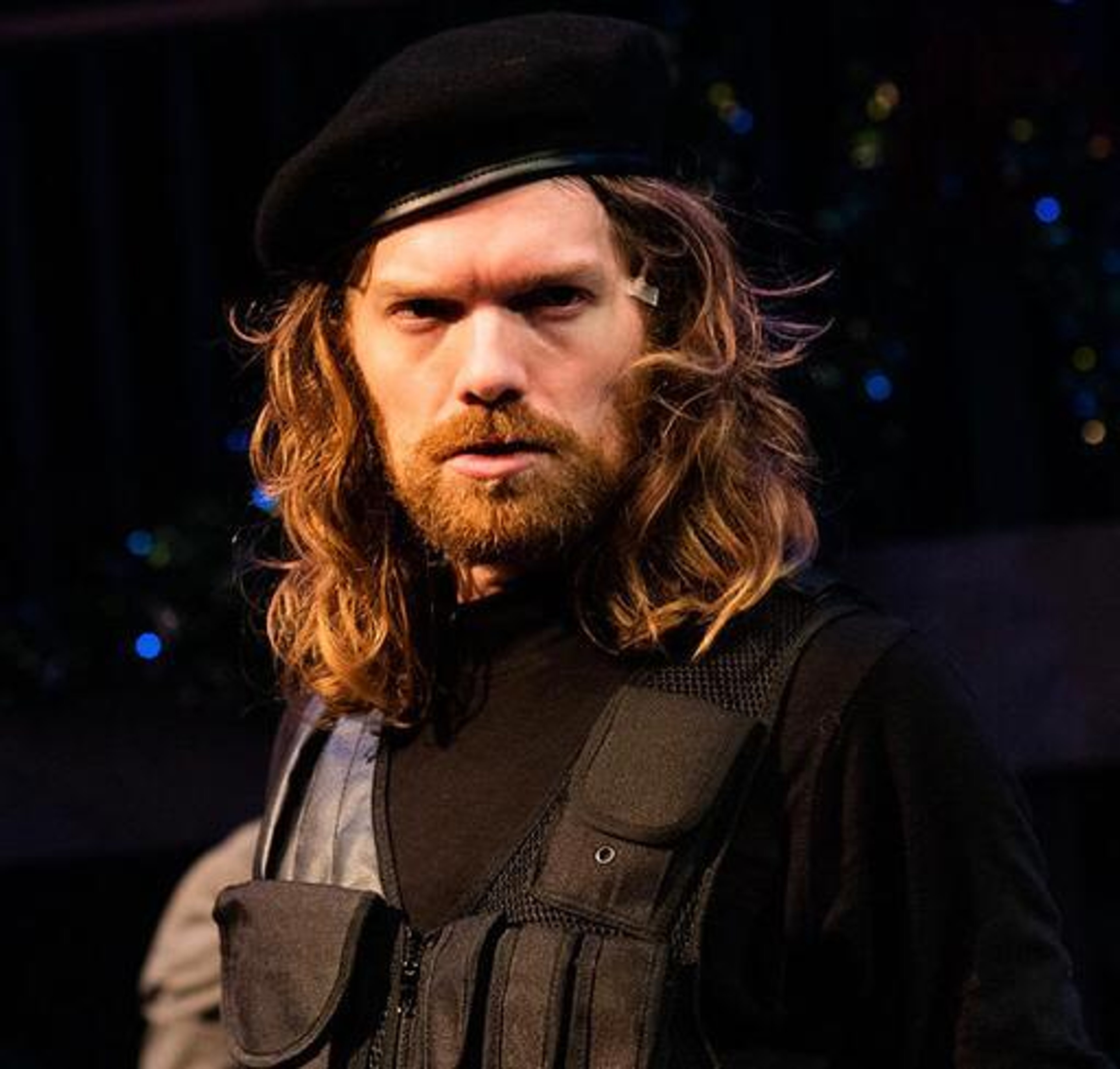A middle-aged man with long hair and a beard wearing a military-style hat and jacket