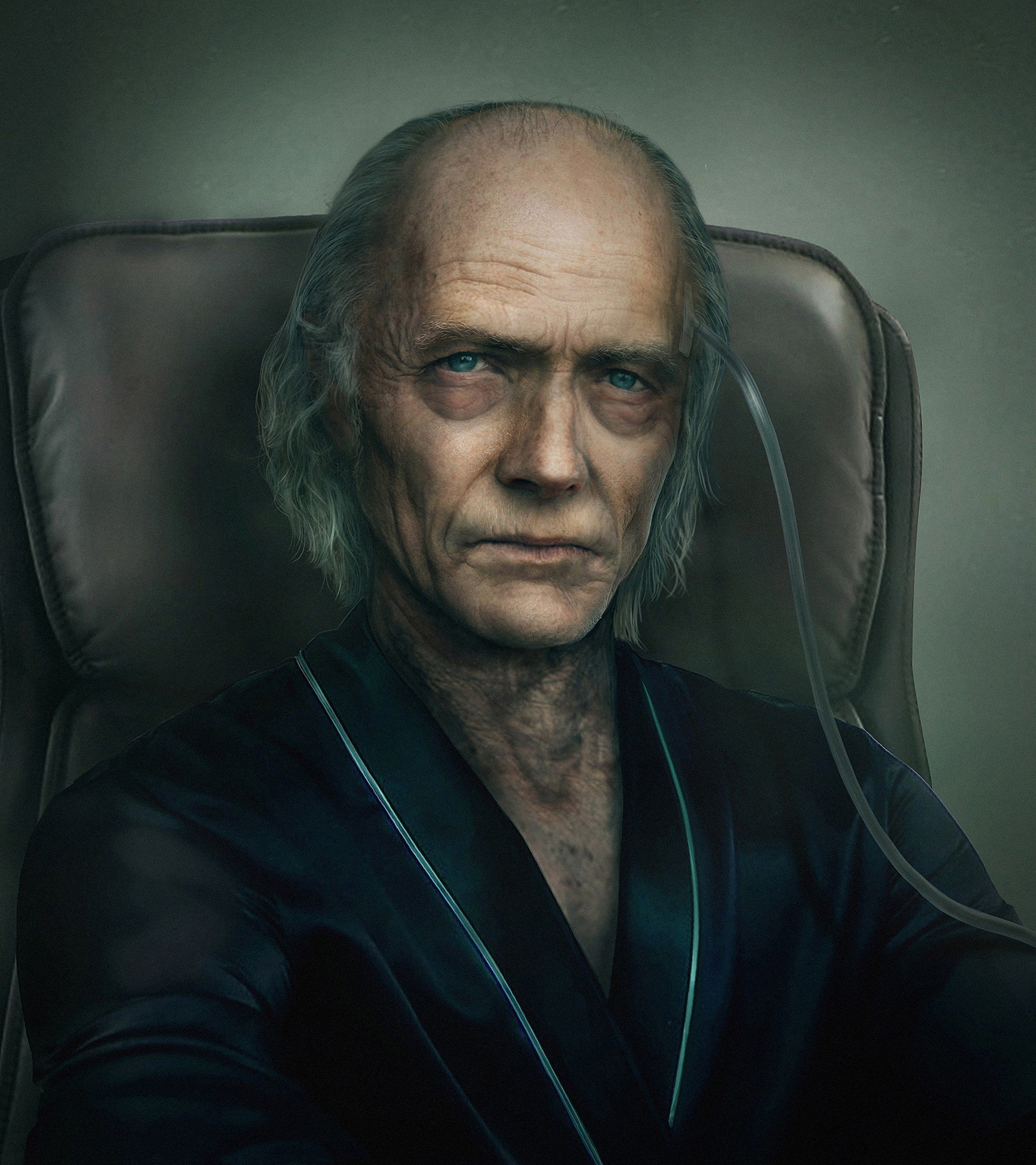 An elderly man with long gray hair wearing a dark suit and sitting in a chair