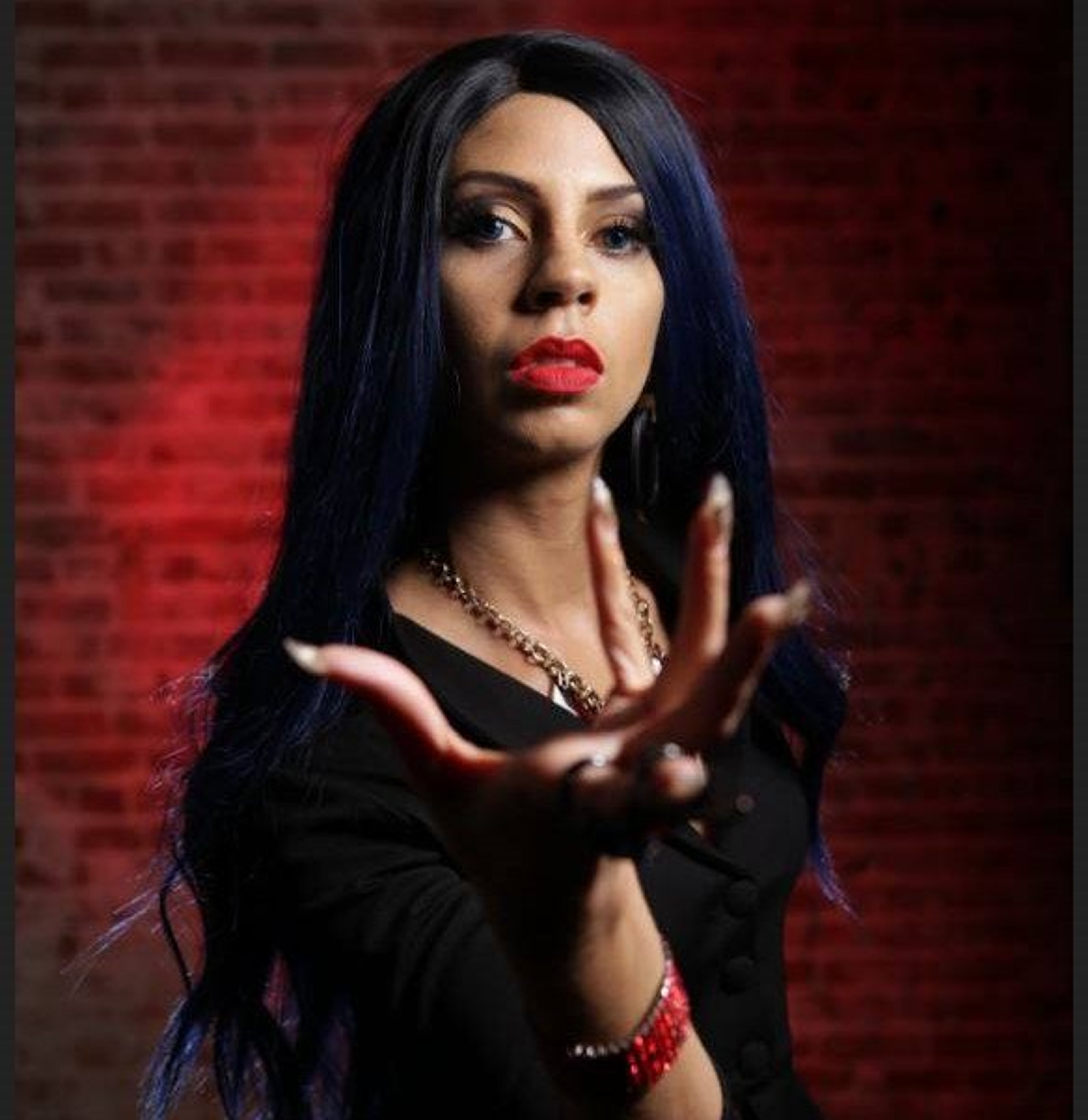 A woman with dark hair wearing a black outfit making a hand gesture against a red background.