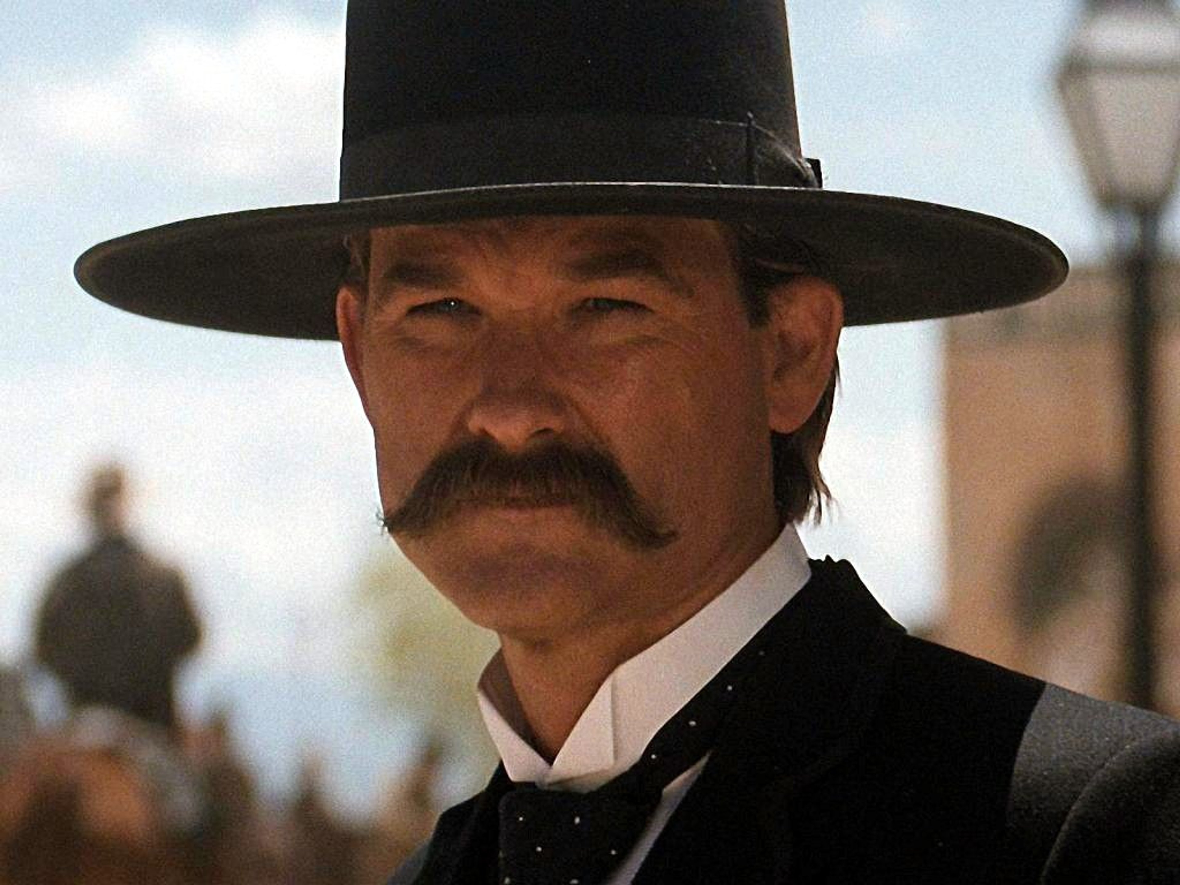 A stern-looking man in a dark suit and hat, standing in a frontier town setting