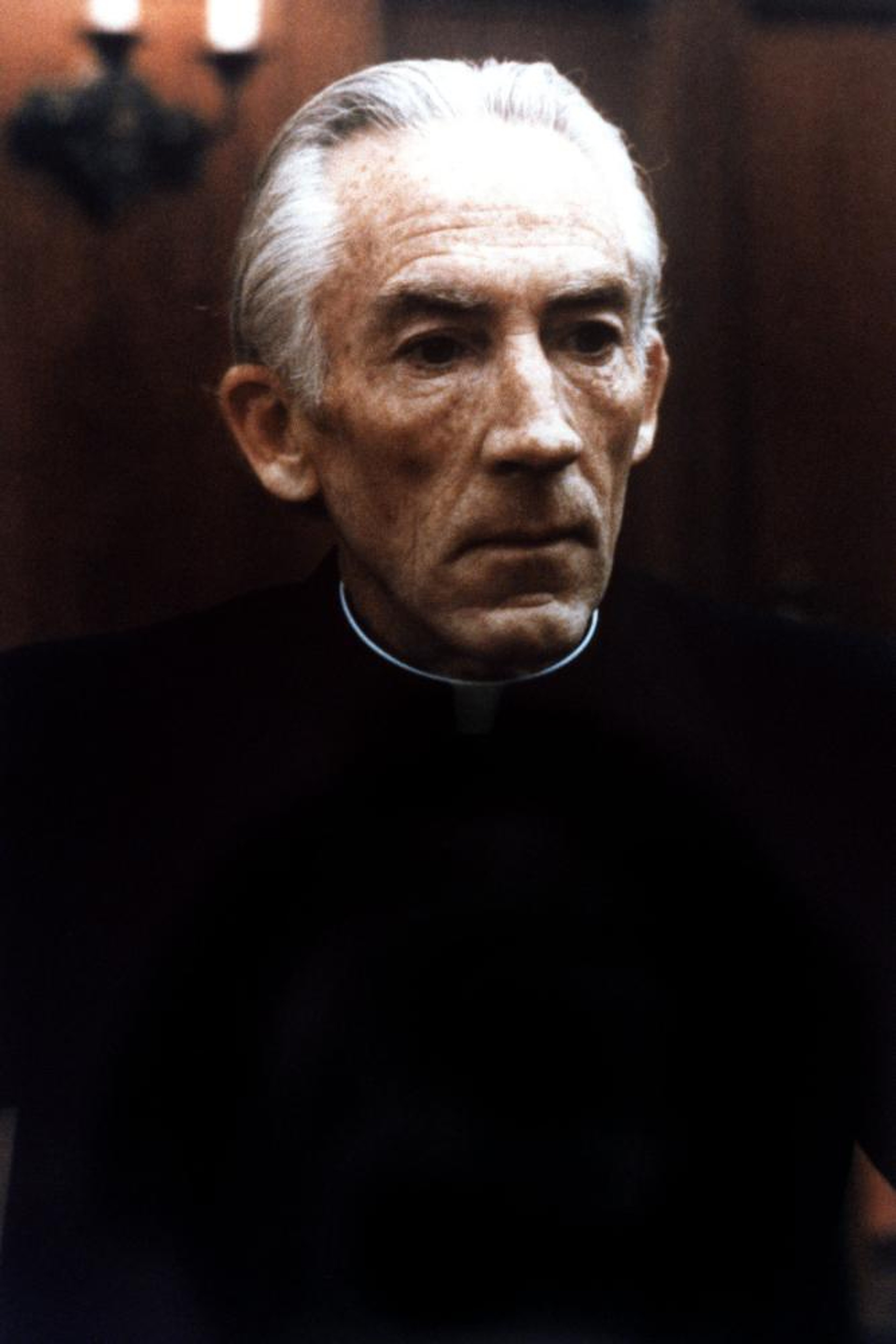 An elderly Catholic priest or religious figure with white hair and a beard, wearing a black clerical robe and having a serious, thoughtful expression.