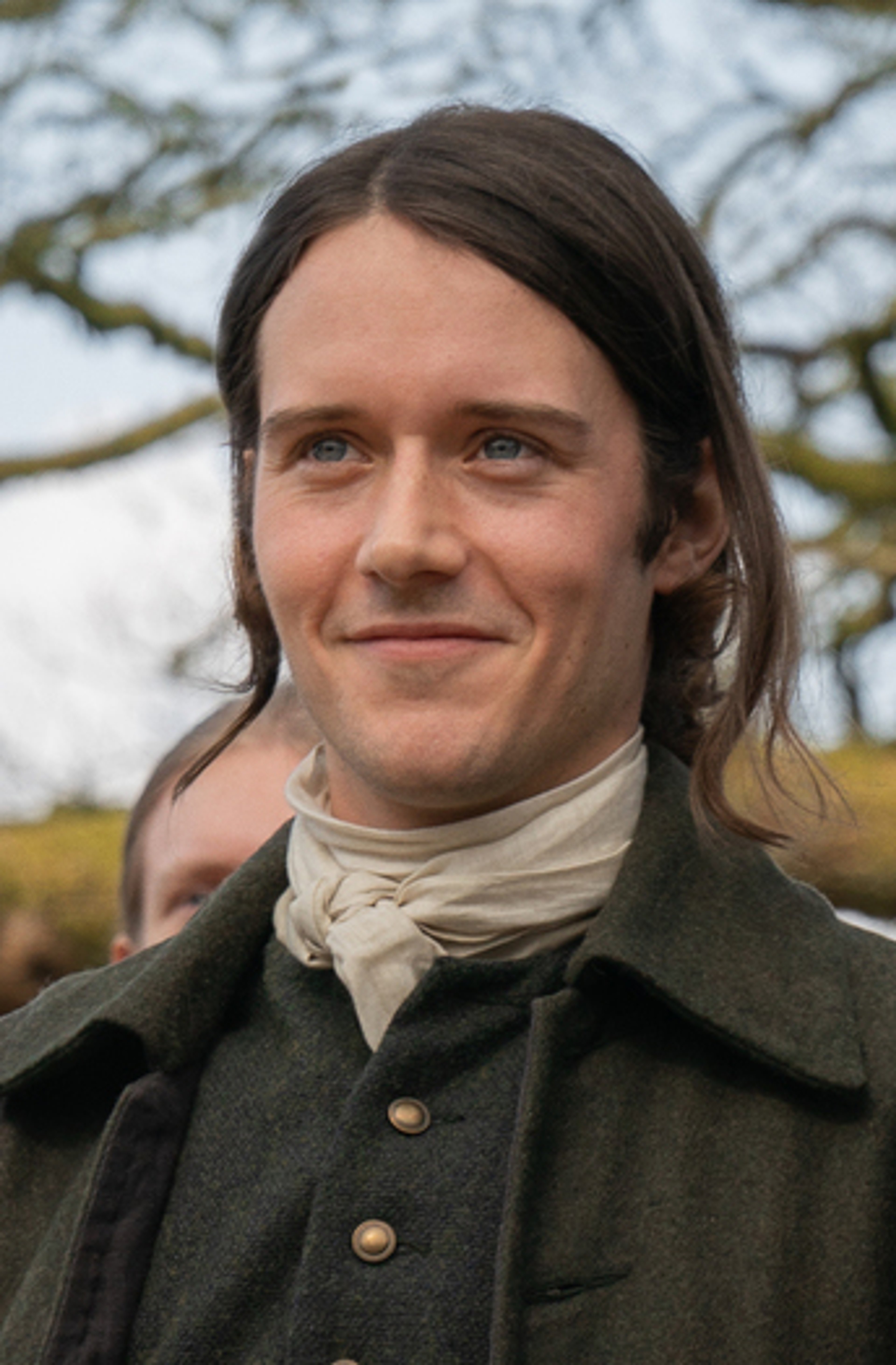 A man with long dark hair and a beard wearing a green coat in an outdoor setting