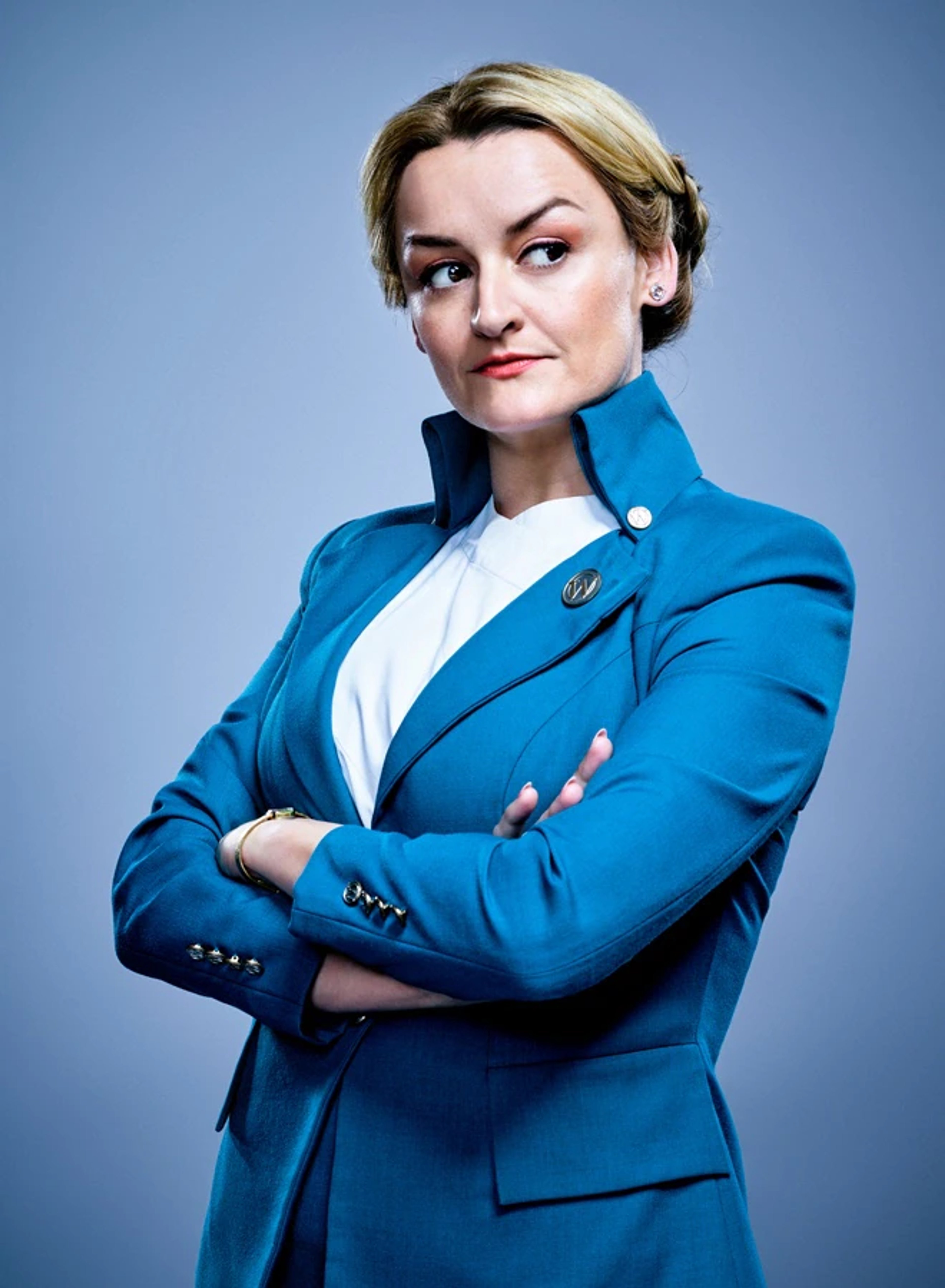 A middle-aged woman with blonde hair wearing a blue blazer and standing with her arms crossed.