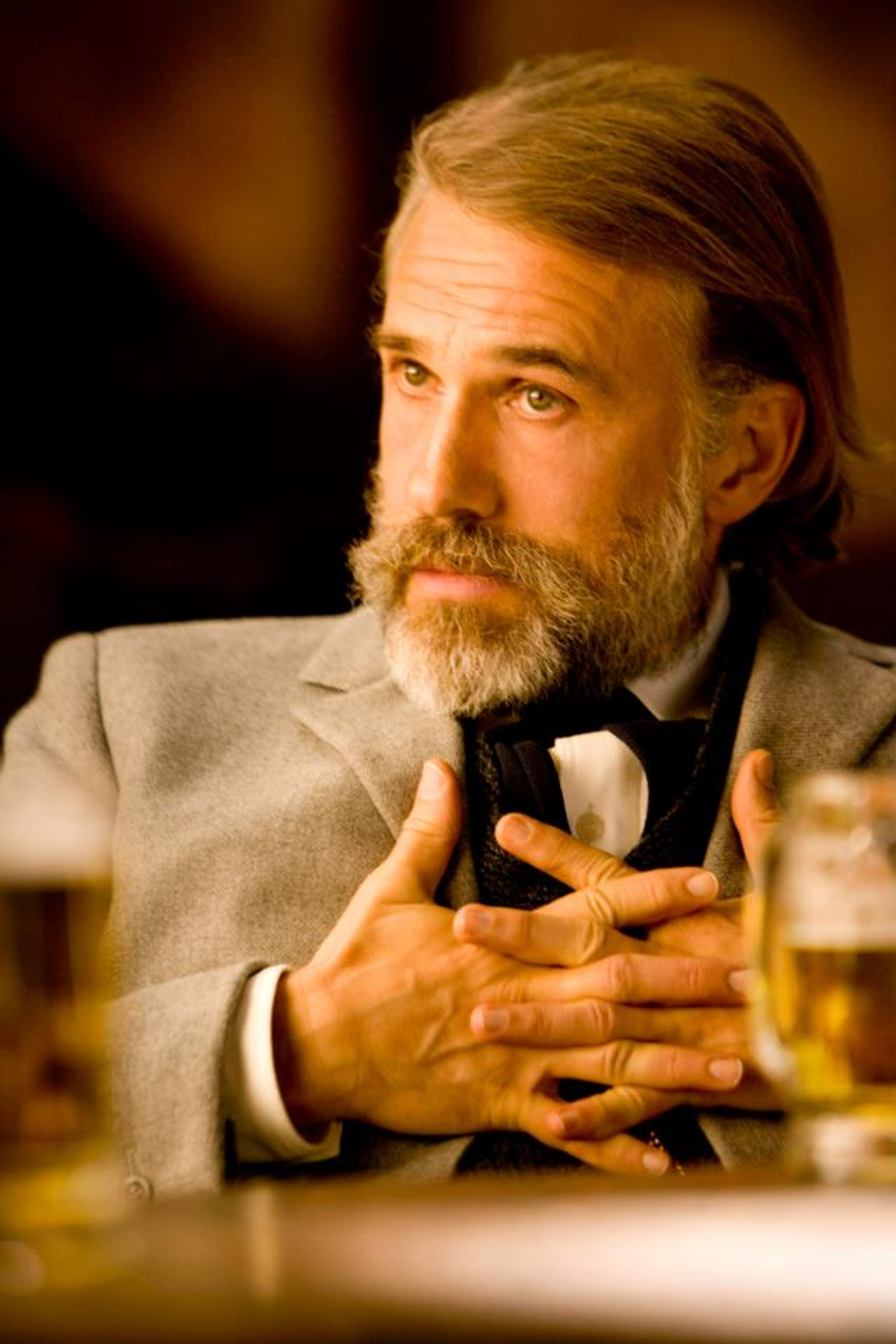 A well-dressed, middle-aged man with a beard and mustache sitting at a table with a glass of alcohol.