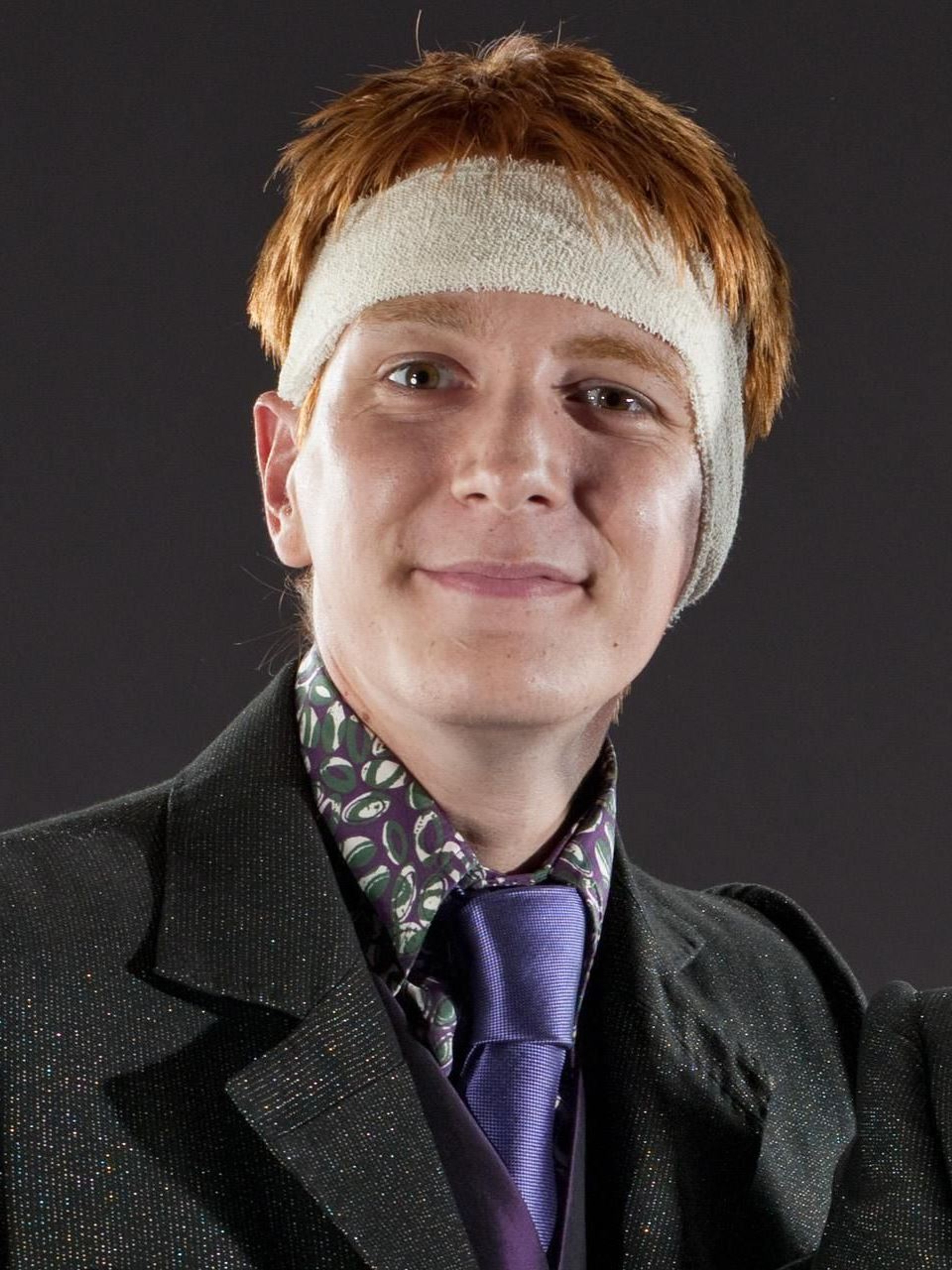 A young man with red hair and freckles, wearing a black coat and a purple tie, with a friendly, mischievous expression.