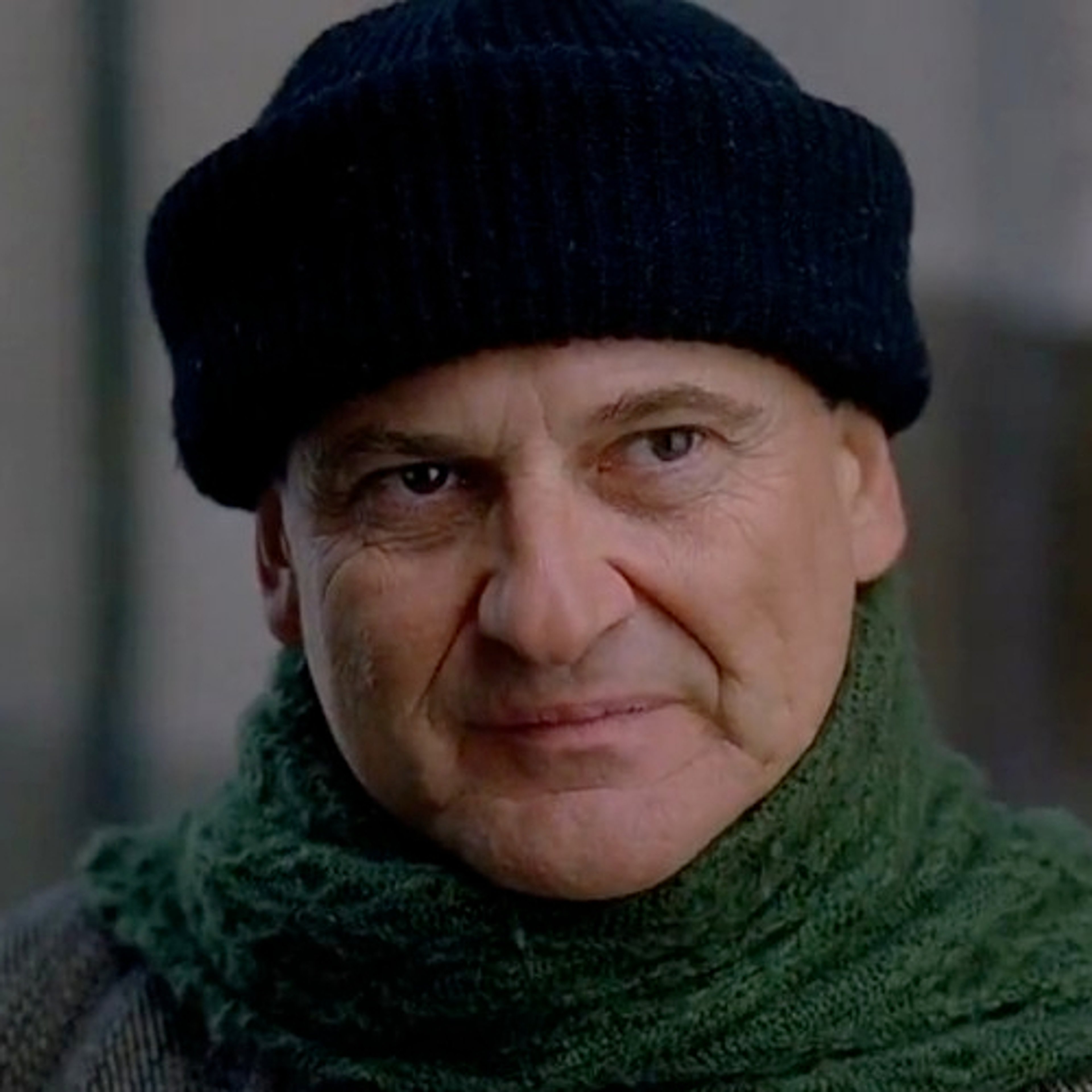 A middle-aged man with a stern expression, wearing a dark beanie hat and green scarf.