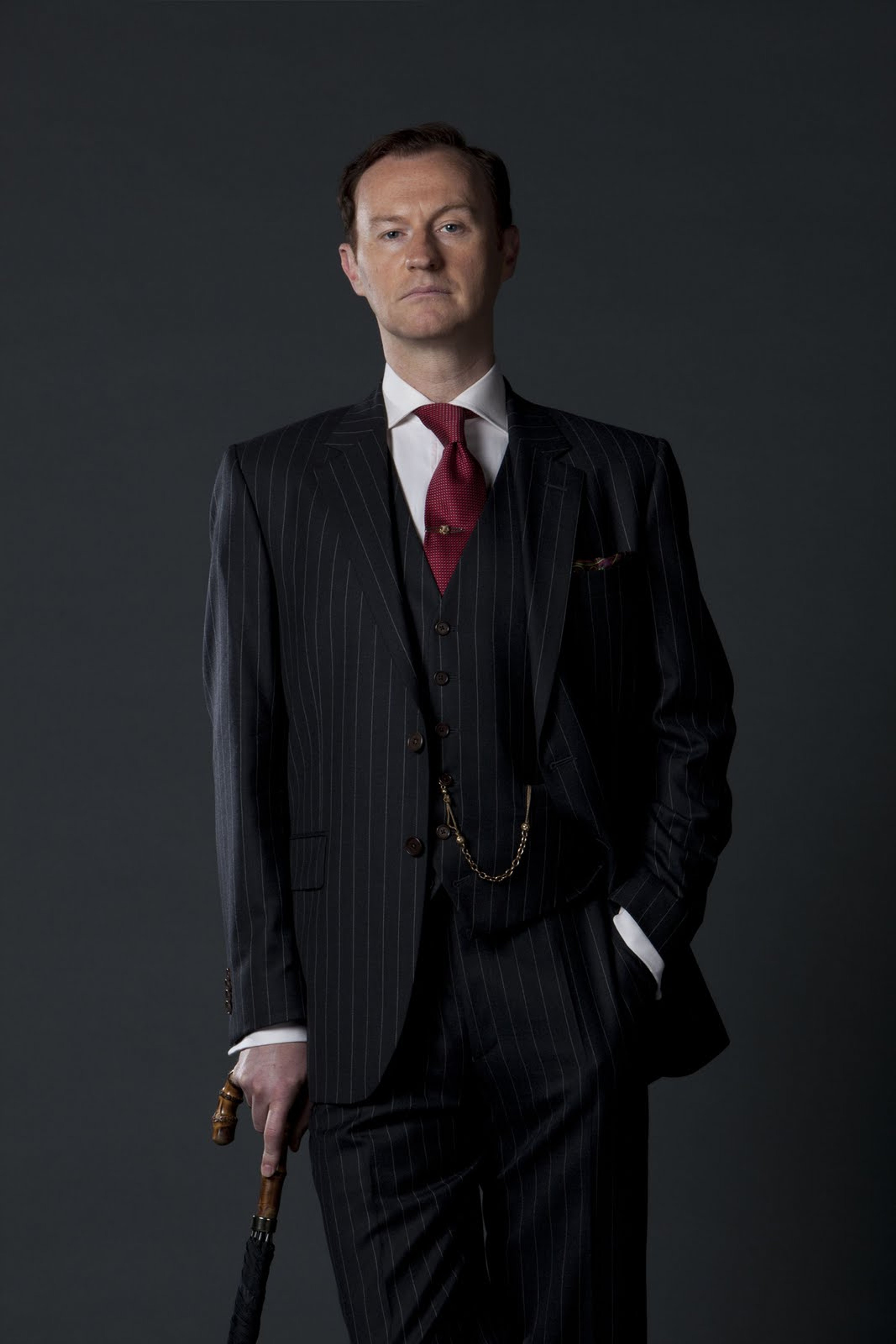 A man in a black suit and red tie standing in front of a dark background