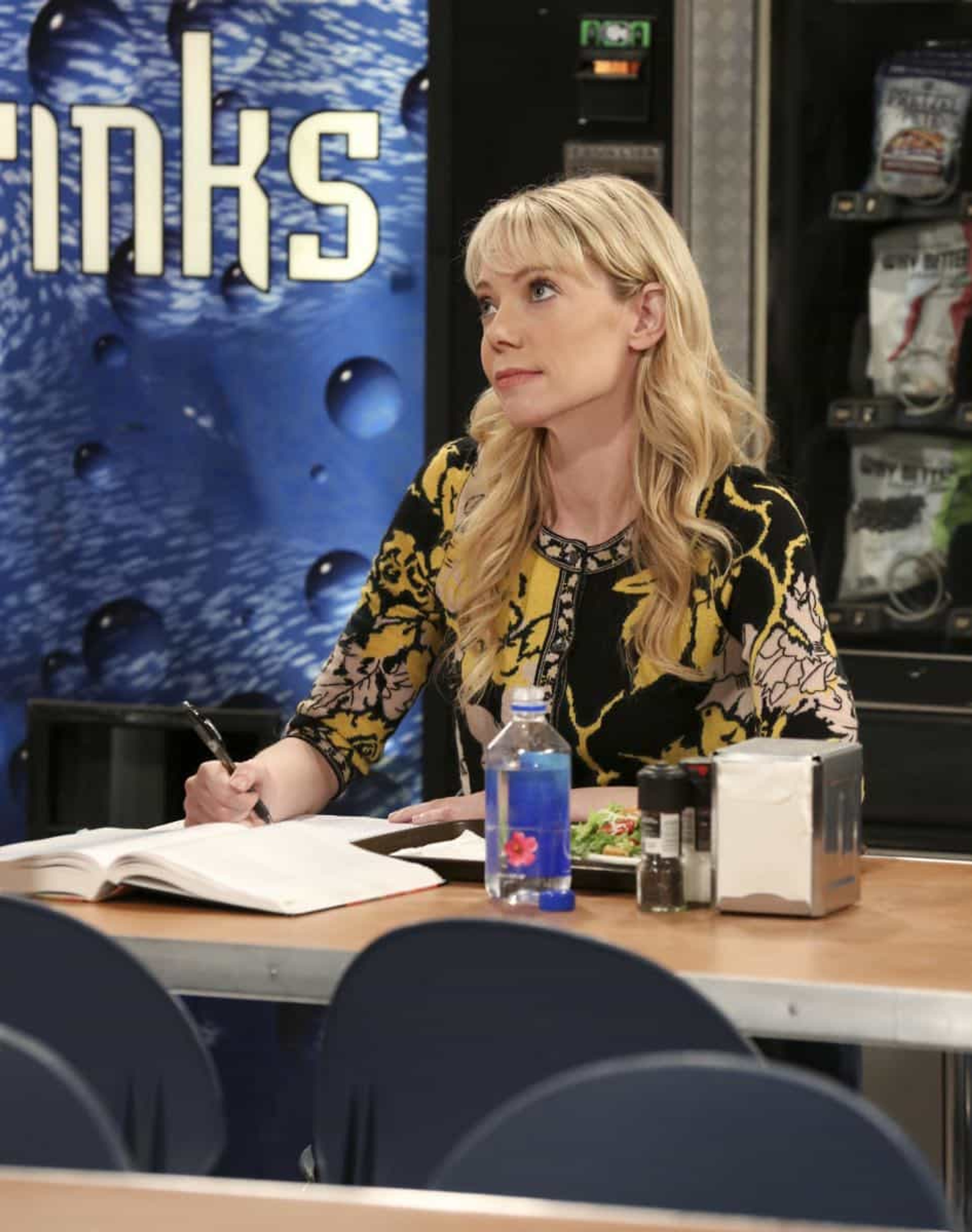 A woman with long blonde hair wearing a patterned dress, sitting at a desk or table with various items in front of her.