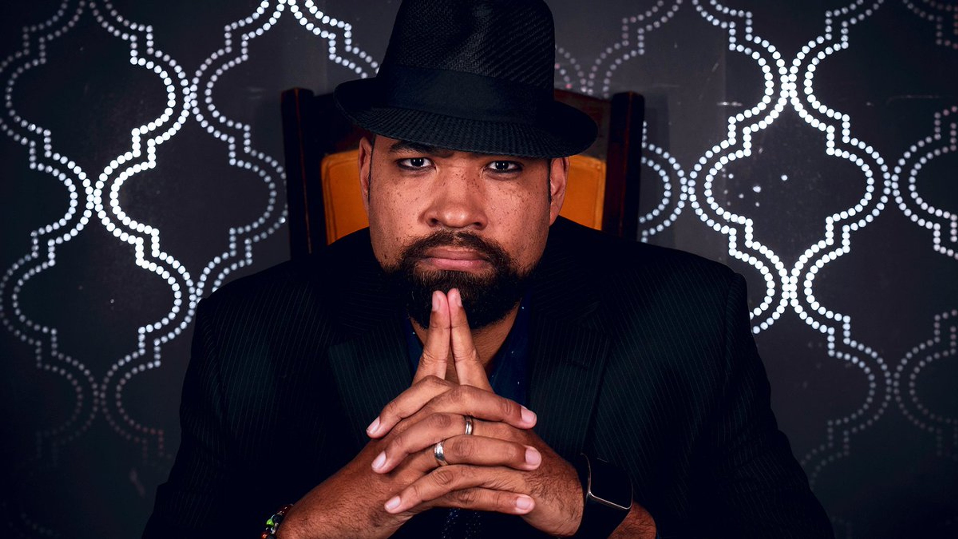 A man in a black suit and hat making a praying or pleading gesture