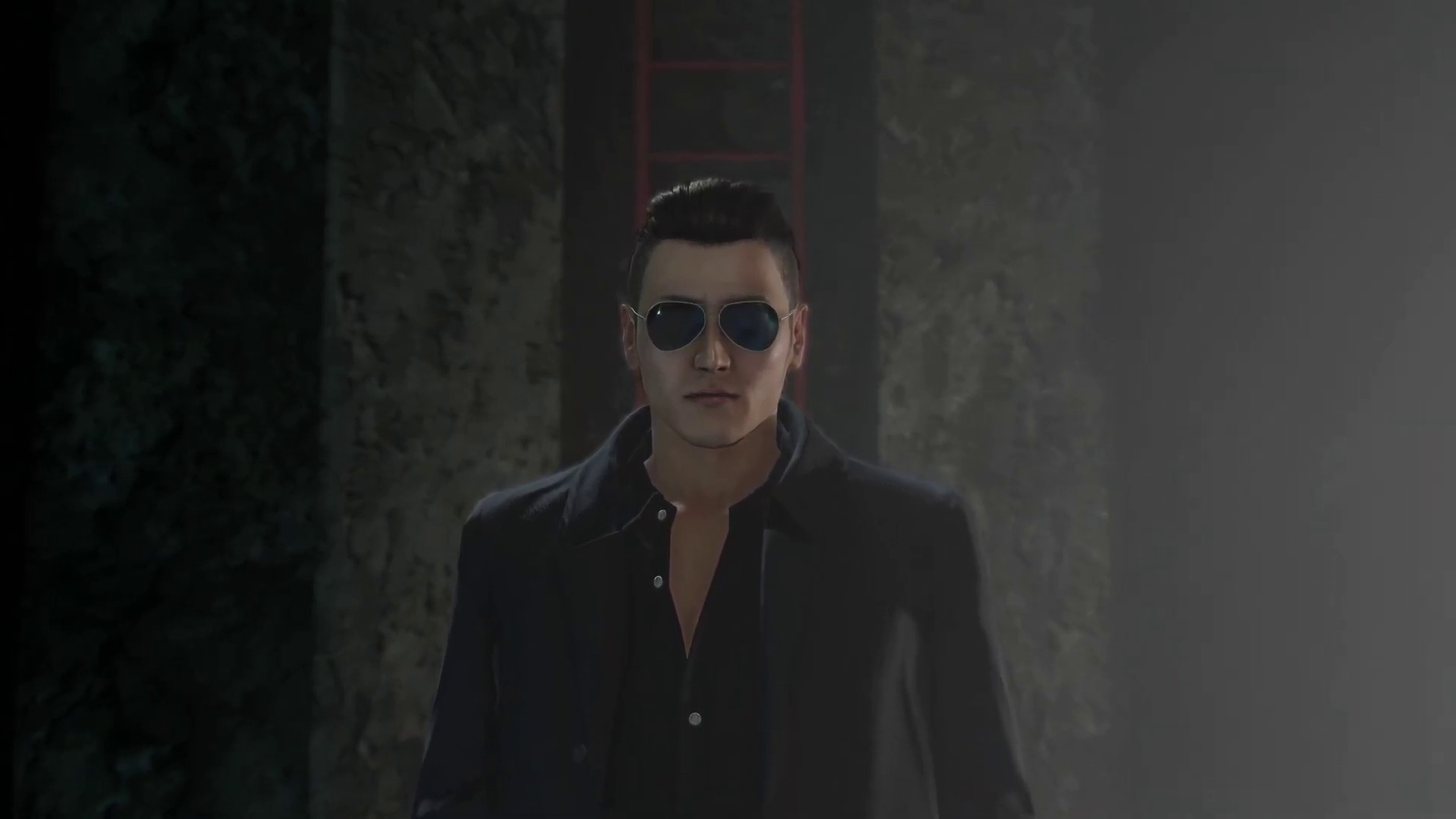 A man with a muscular build, slicked-back black hair, and sunglasses, wearing a black suit and scowling intensely.