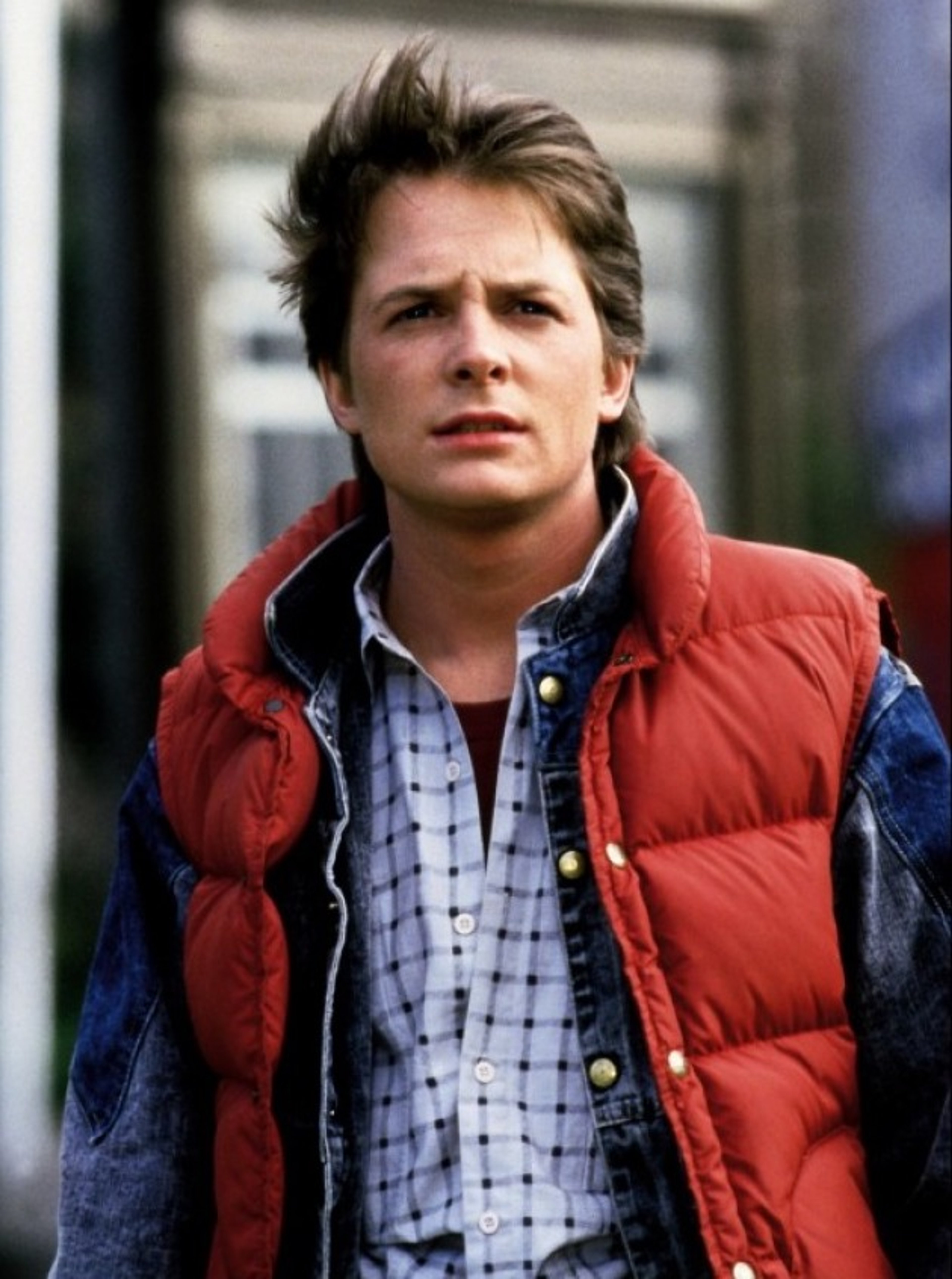 A young man with short brown hair wearing a red jacket, plaid shirt, and blue jeans