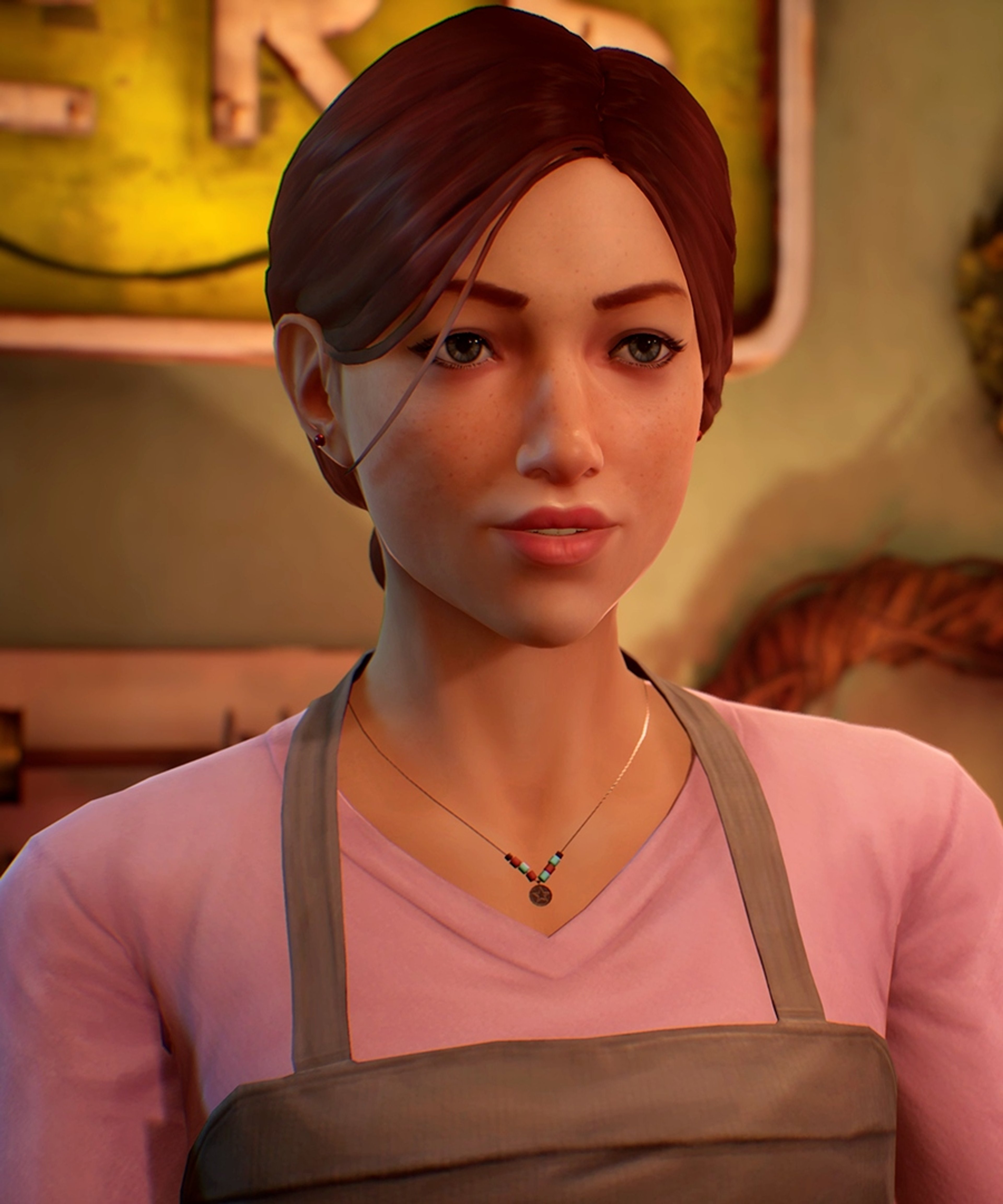 A young woman with dark hair and a friendly expression, wearing a pink shirt in a cozy indoor setting.