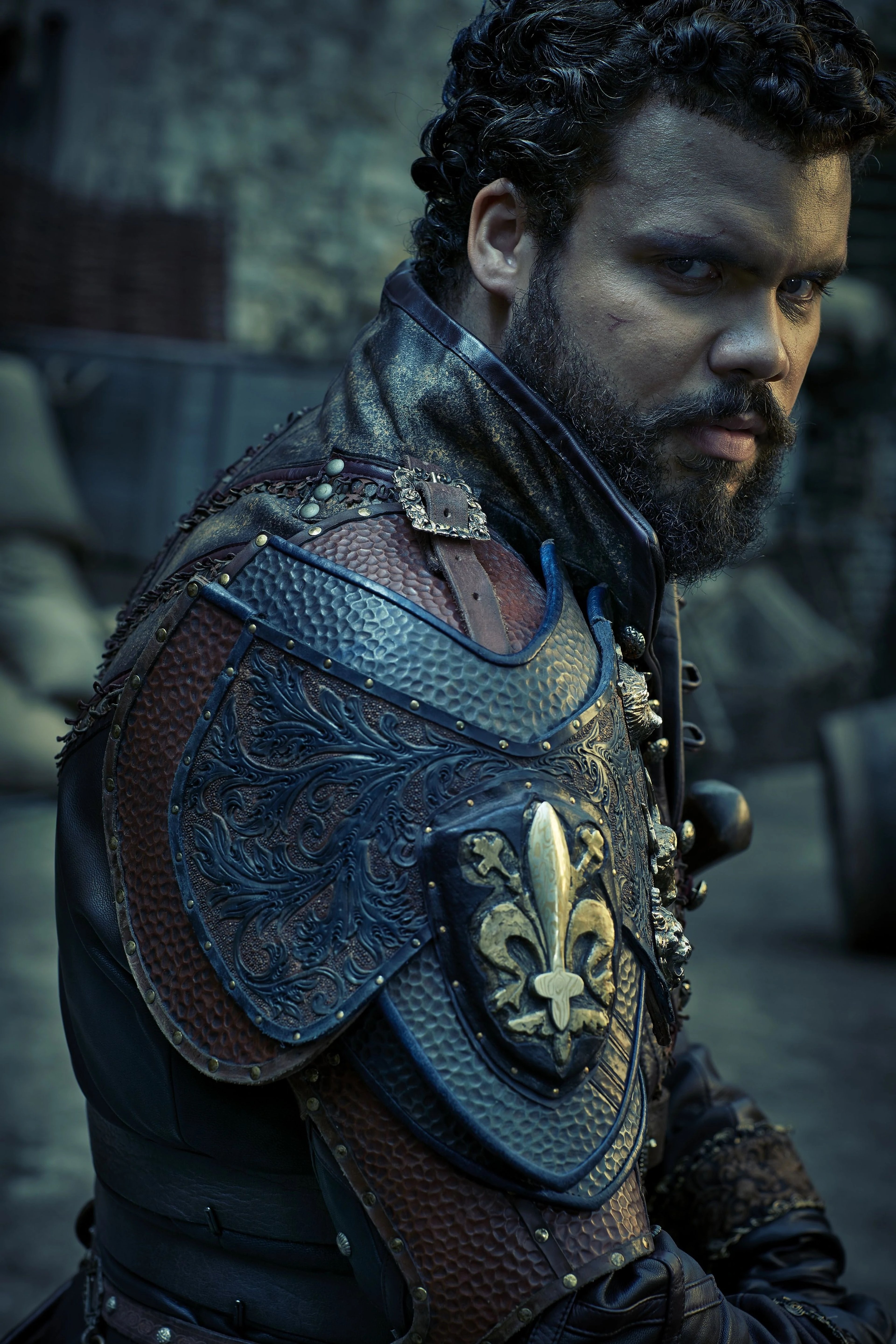 A man with a muscular build wearing a blue and gold doublet and cloak, with a stern expression on his face.