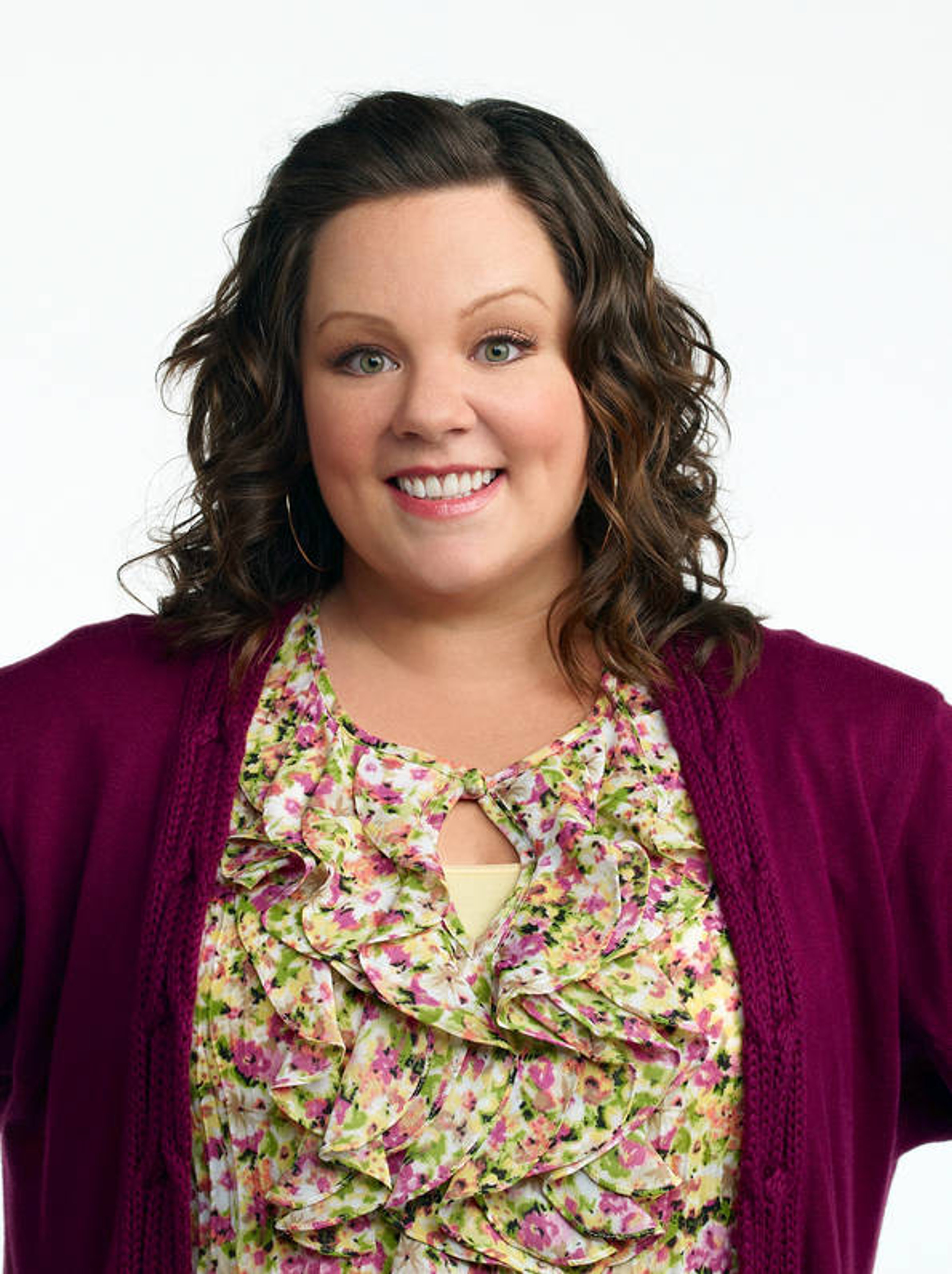 A smiling woman with curly brown hair wearing a floral top and purple cardigan