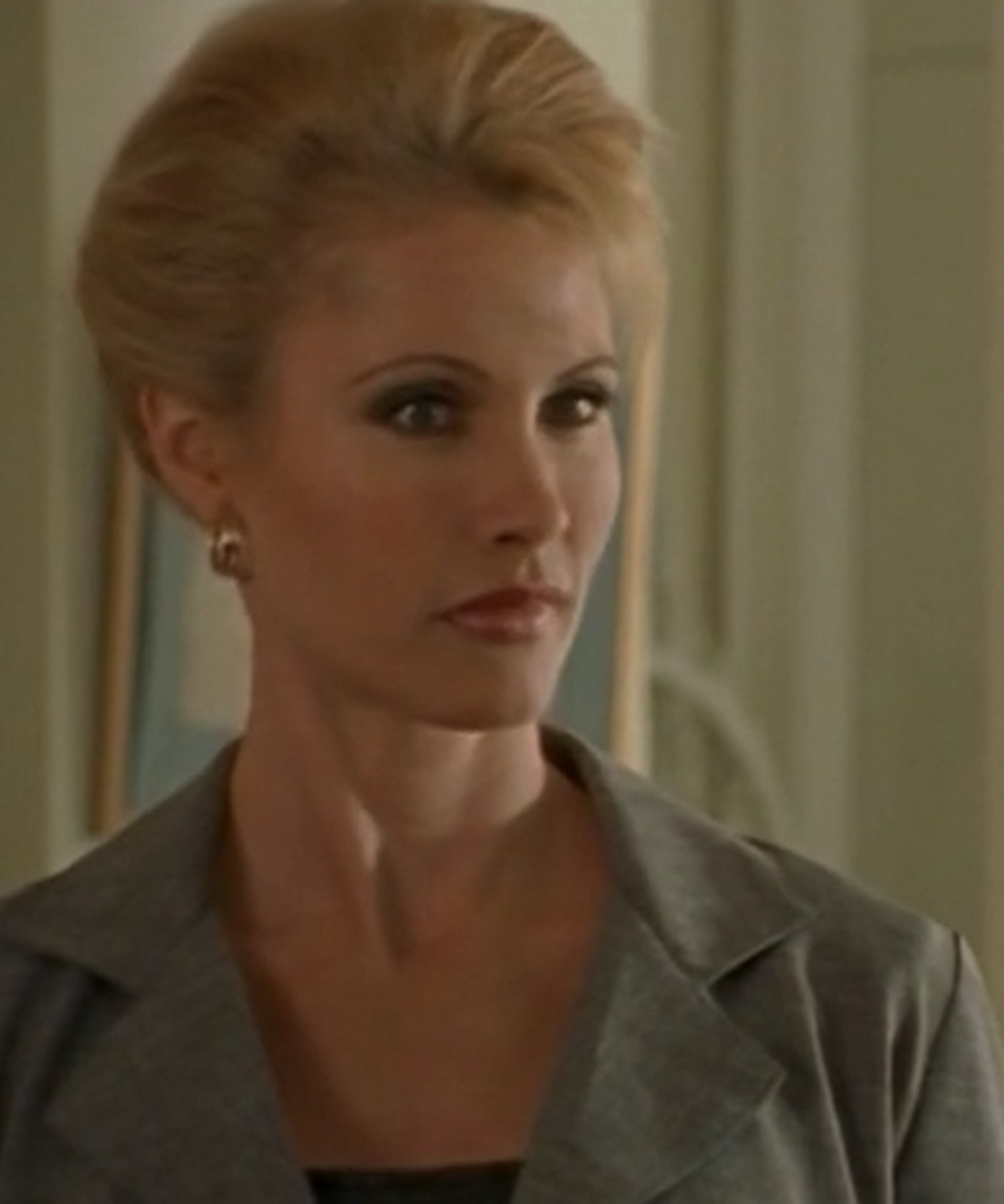 A woman with blonde hair wearing a gray blazer, looking serious.