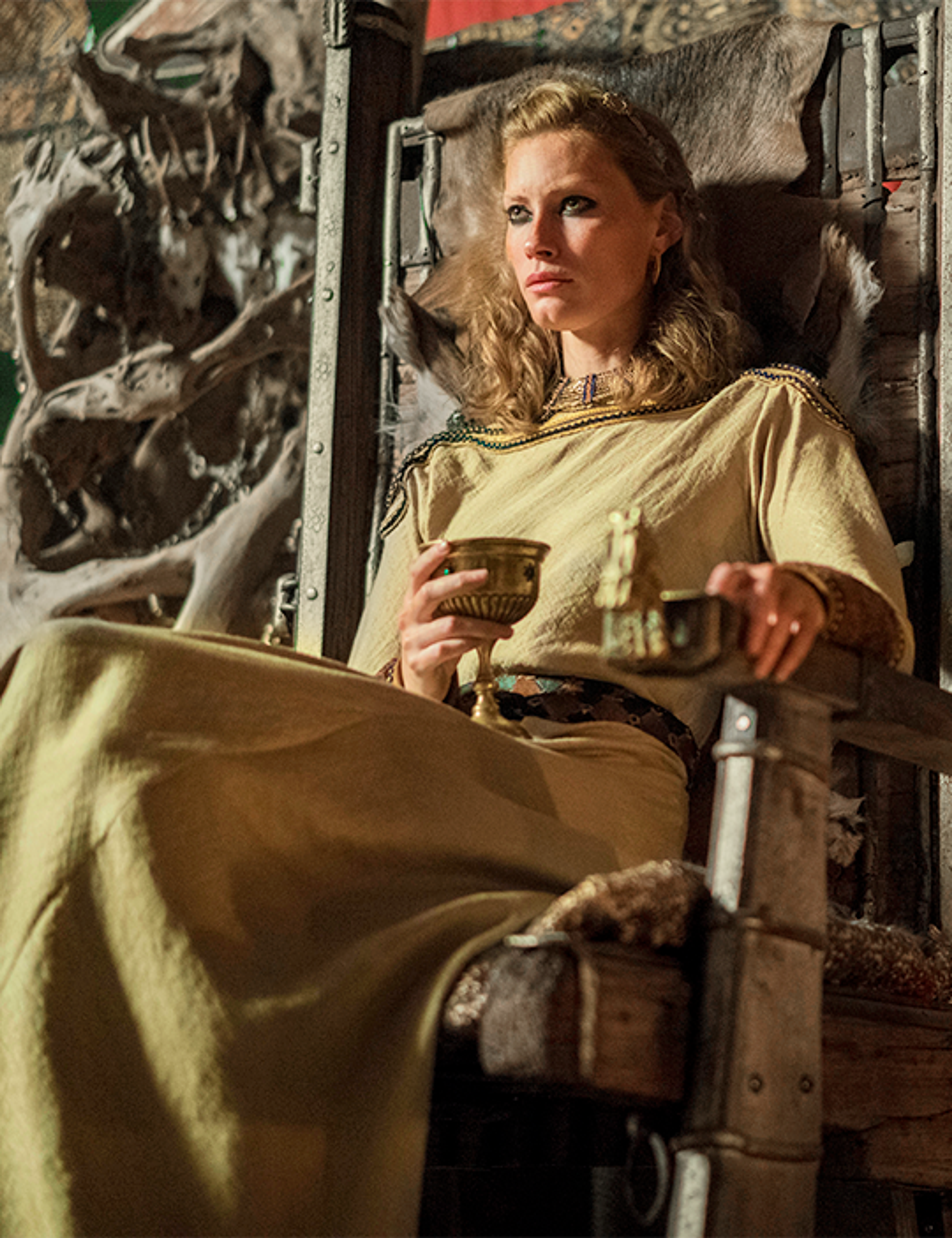 A woman with long blonde hair sitting in a rustic, medieval setting, holding a cup or mug.